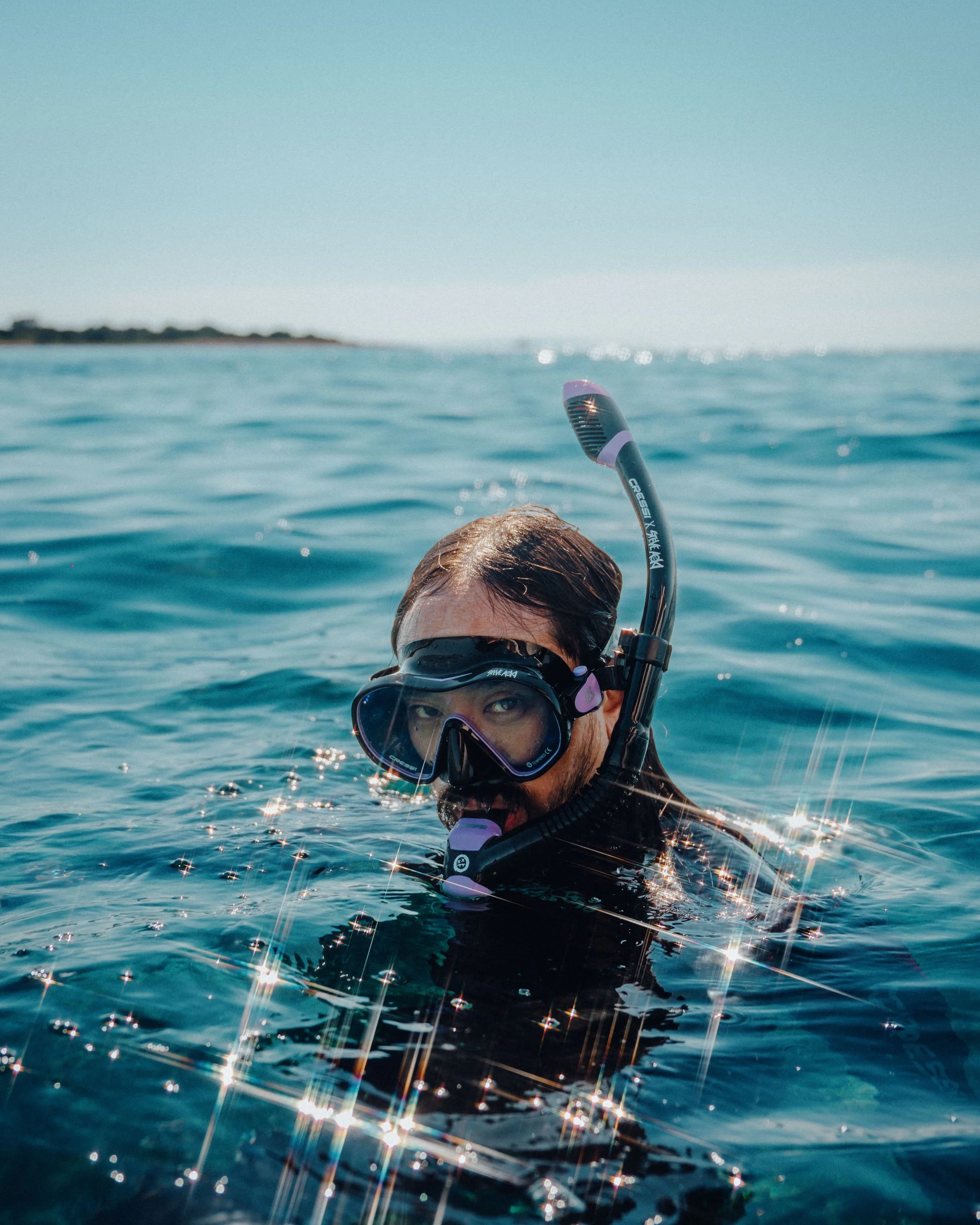 Steve Aoki wearing Cressi Diving equipment in a wetsuit with a snorkel and mask swimming in the ocean on a sunny day.