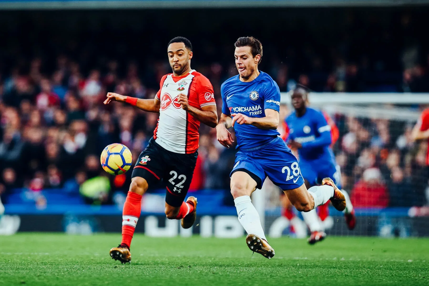 Two soccer players competing for the ball during a match, with a crowd in the background.
