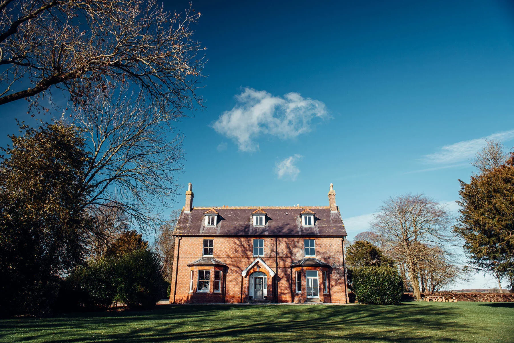 Abbots Court Rural Retreat, Dorset. A large brick house with three stories, a steep roof, and dormer windows, surrounded by trees and a well-maintained lawn under a blue sky with scattered clouds.