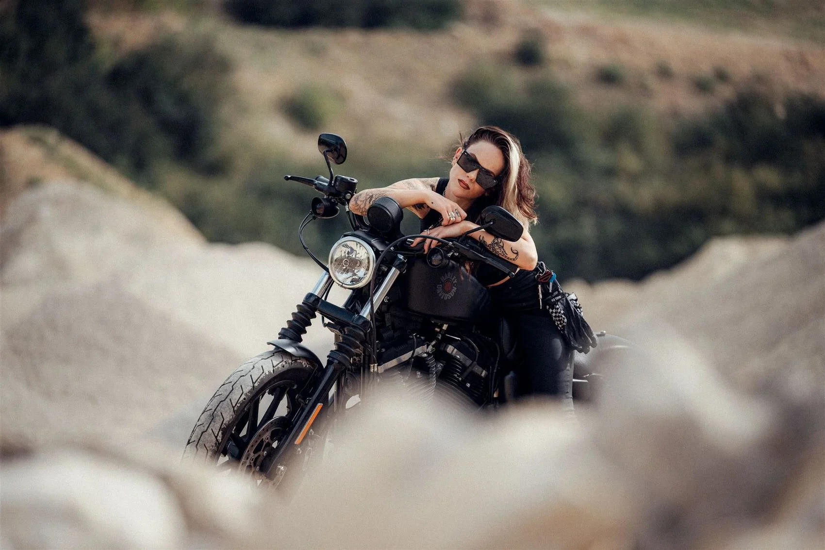 A woman with dark hair wearing sunglasses and black clothing is leaning on a black motorcycle in a desert landscape with rocky terrain and hills in the background.