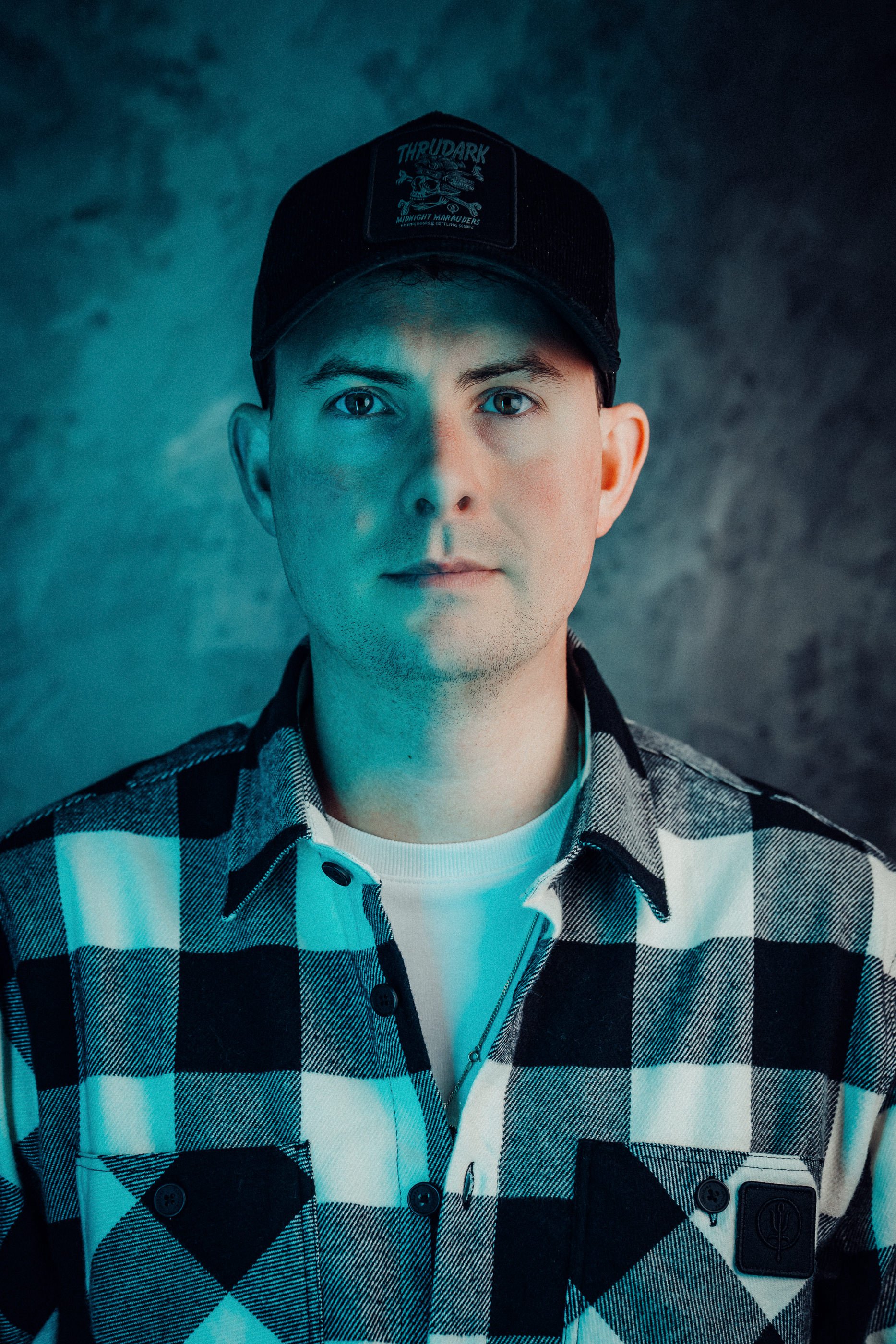 James Bridle Photography Headshot, Face on. wearing a check shirt and Black Thrudark cap.