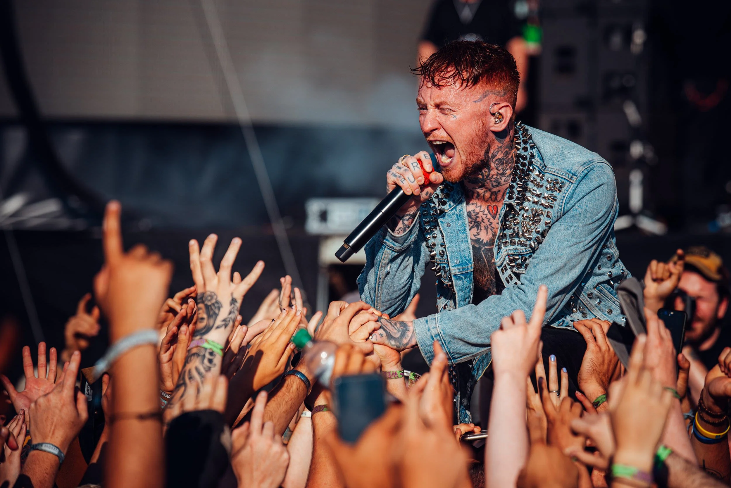 Male singer with tattoos, wearing a denim jacket with studs, performing at a concert, singing into a microphone and engaging with the audience.