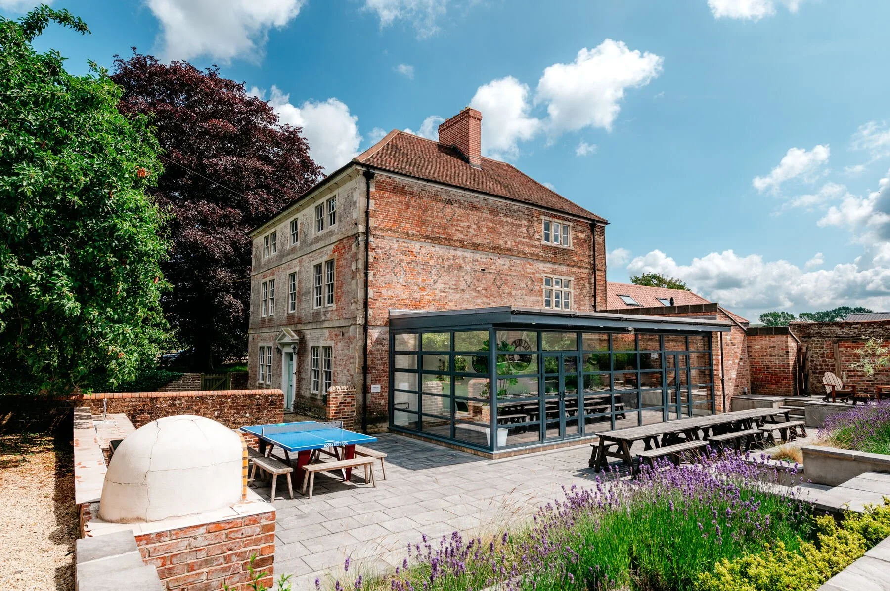 Interior and Exterior Photography of outdoor patio with a brick house, a glass enclosed structure, picnic tables, a ping pong table, and purple flowering plants, under a partly cloudy sky.