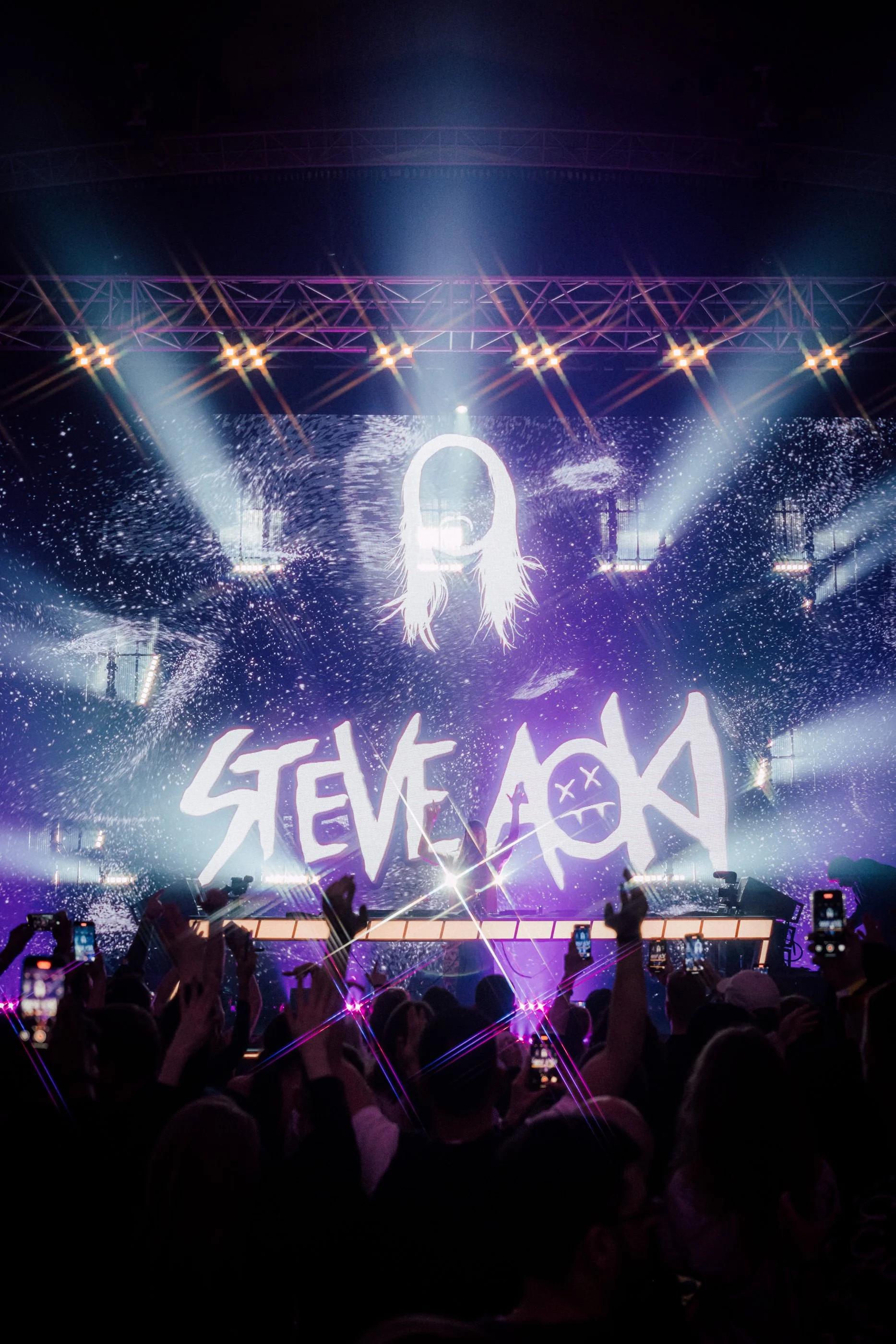 Steve Aoki playing a private event in Barcelona scene shows stage with bright lights and a large screen displaying a stylised mans face with the name 'Steve Aoki' in bold letters.