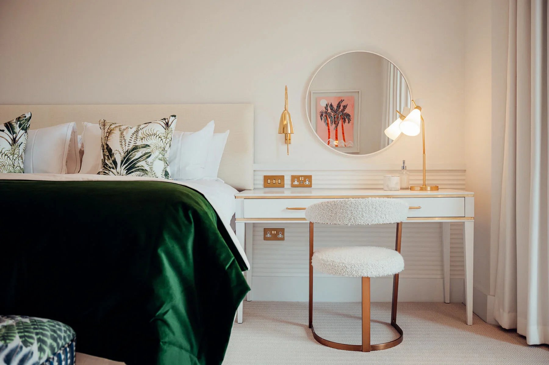 A bedroom with a white bed, pillows with leaf designs, a white desk with gold accents, a round mirror, a modern white and gold chair, and a side lamp. Neutral tones with green, gold, and white accents.