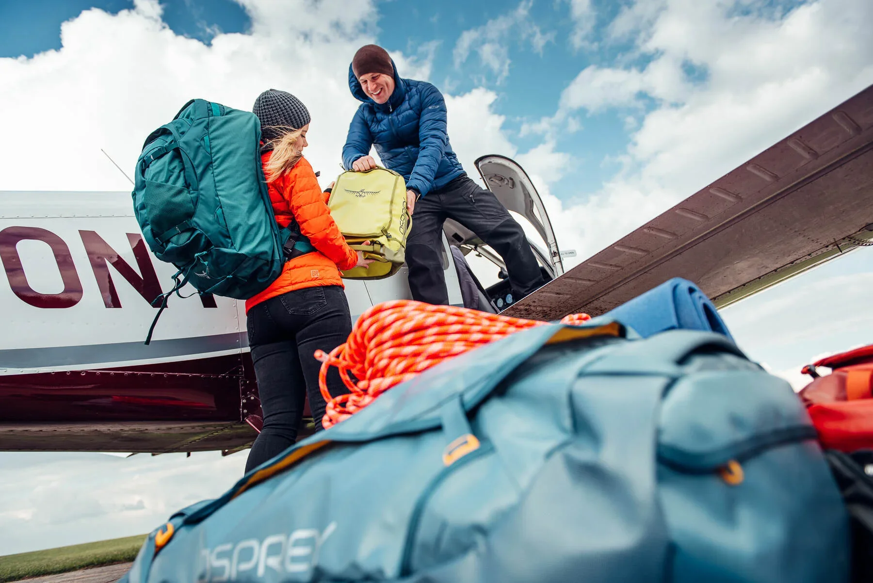 Shot for Osprey Europe - A man and a woman are loading luggage onto a small airplane with a step ladder. The woman is wearing a red jacket and carrying a large teal backpack, and the man is wearing a blue jacket and a brown beanie. 