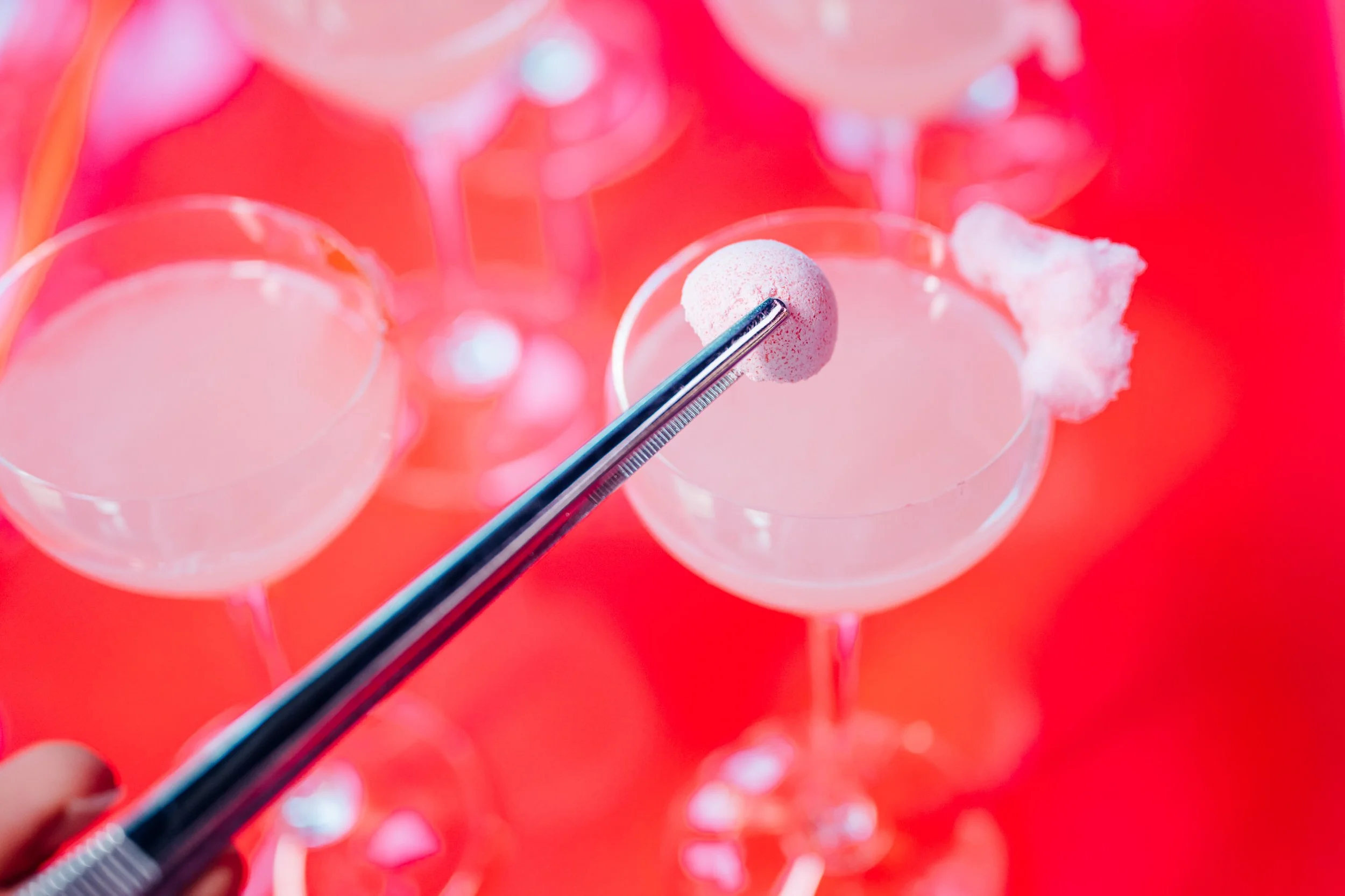 LUSH Products in use at Oxford Street Store Halloween 2023. A hand holding tweezers with a pink macaron over two empty cocktail glasses with cotton candy garnishes, set against a pink background. Captured by James Bridle Event Photographer London. 