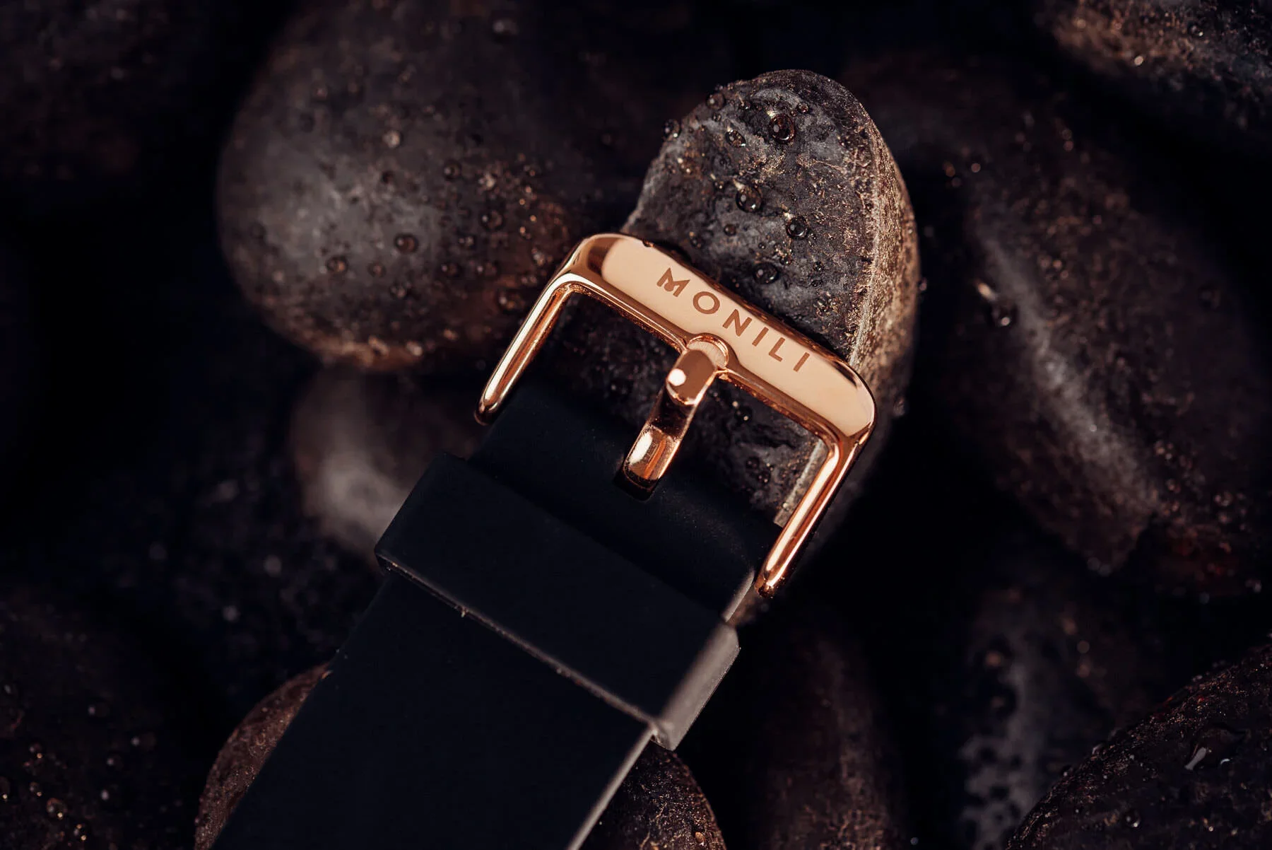 Close-up of a black watch strap with a gold buckle engraved with 'MONILI', resting on dark volcanic rocks.