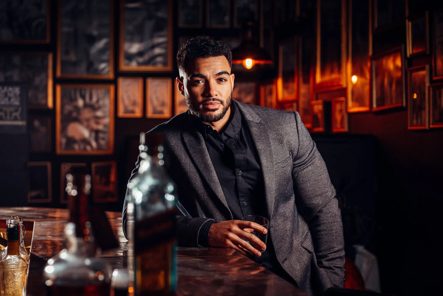 A man in a gray blazer and black shirt sitting at a bar with bottles in the foreground, in a dimly lit bar or club with framed pictures on the wall behind him.