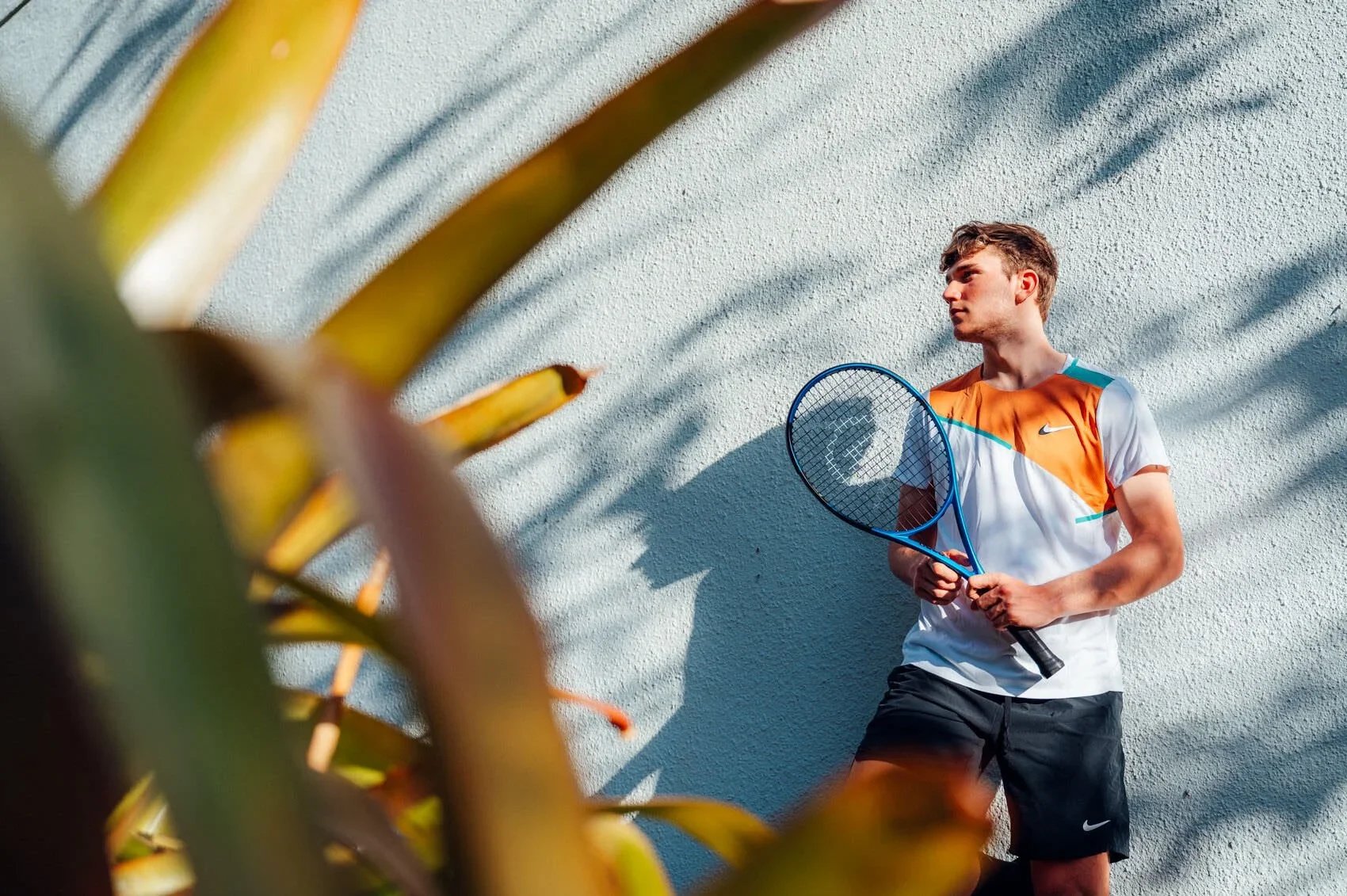 Young man in an orange and white Nike sports shirt standing with tennis racket against a textured white wall, casting shadows from nearby plants.