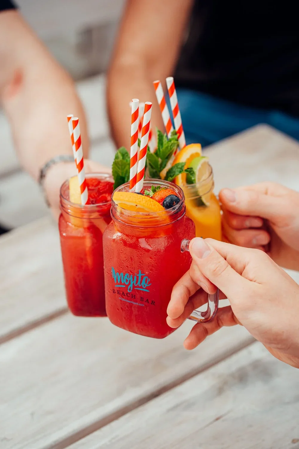 Three mason jars filled with colourful fruit drinks, garnished with fruit slices, mint, and striped straws. One hand is holding the jar labeled "Mojito BEACH BAR," and three people are reaching for glasses.