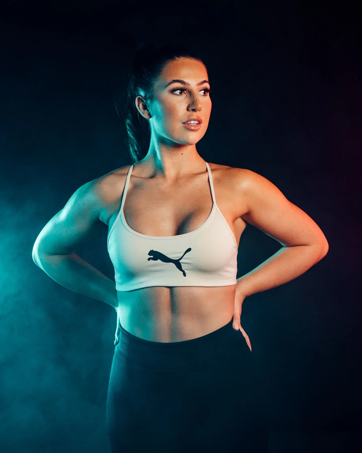 Fitness Photoshoot by James Bridle Photography UK - A woman with dark hair in a ponytail wearing a white sports bra with a Puma logo and black workout pants, standing with her hands on her hips against a dark background with colorful lighting.