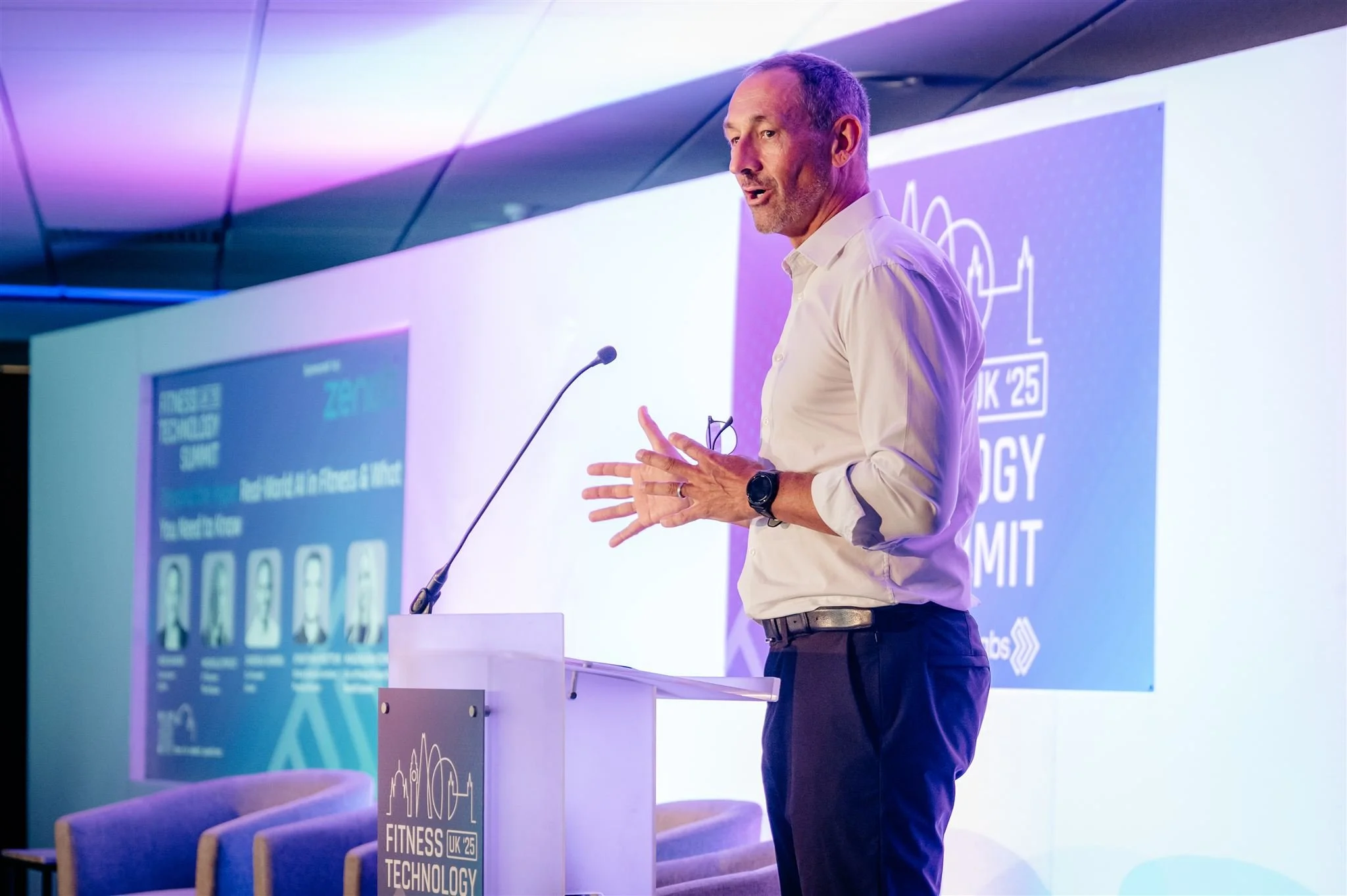 Fitness Technology Summit at Stamford Bridge, London. A man giving a presentation at a conference, standing behind a podium with a microphone, with a large screen displaying event information in the background. Captured by James Bridle Photography.