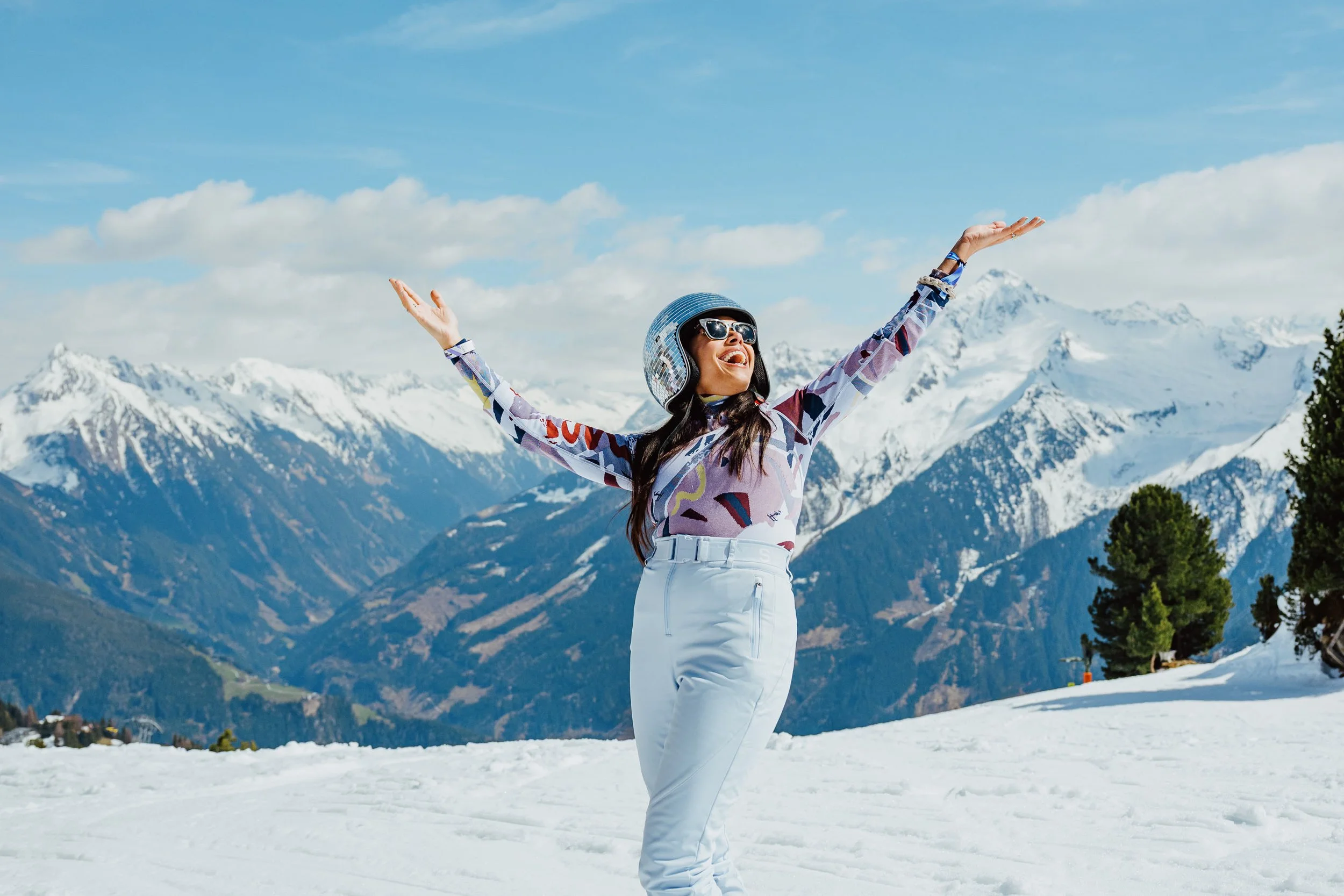 Snowbombing 2025 Woman in winter clothing and helmet with arms spread wide, smiling, standing on snowy ground with snow-capped mountains and blue sky in the background.
