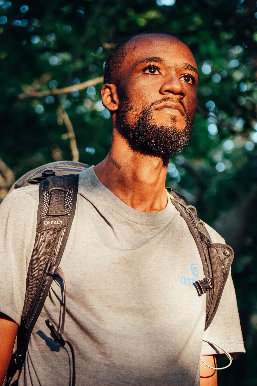A man with a beard, wearing a gray t-shirt and a backpack, standing outdoors in a wooded area during the daytime.