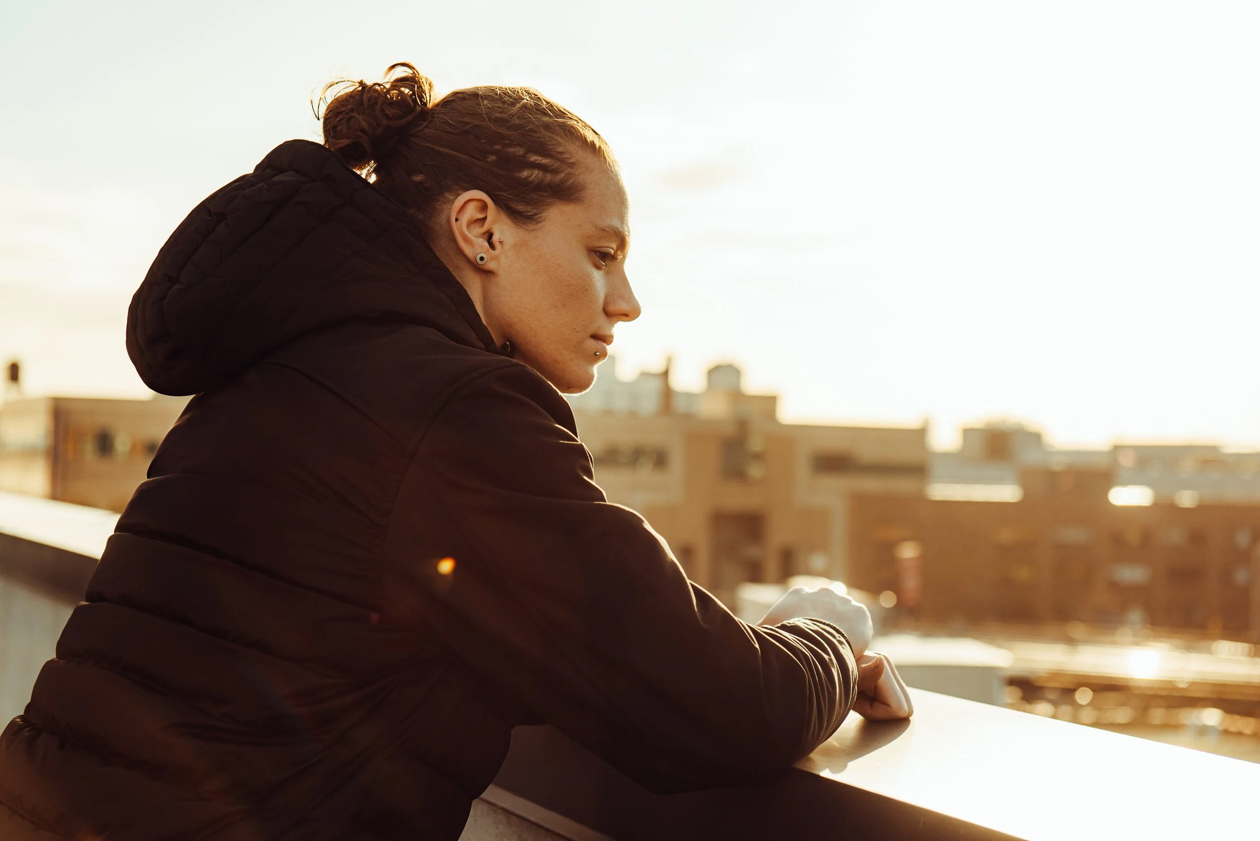 Dunlop Sports Commercial Shoot in NYC, USA. A Young woman with curly hair, wearing a black jacket, looking down while standing on a balcony at sunset with city buildings in the background.