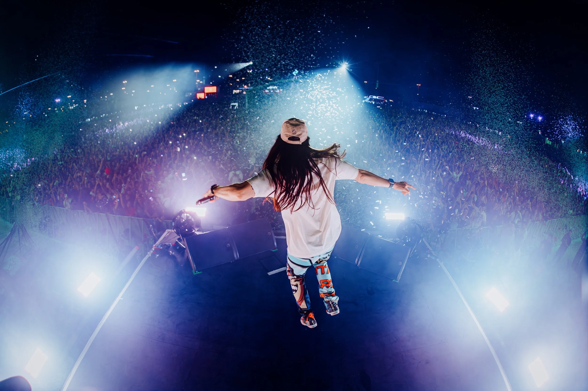 Steve Aoki on stage in Dubai UnTold Festival with his back to the camera, arms outstretched, facing a large crowd at a concert or festival with colorful lights and confetti in the air.