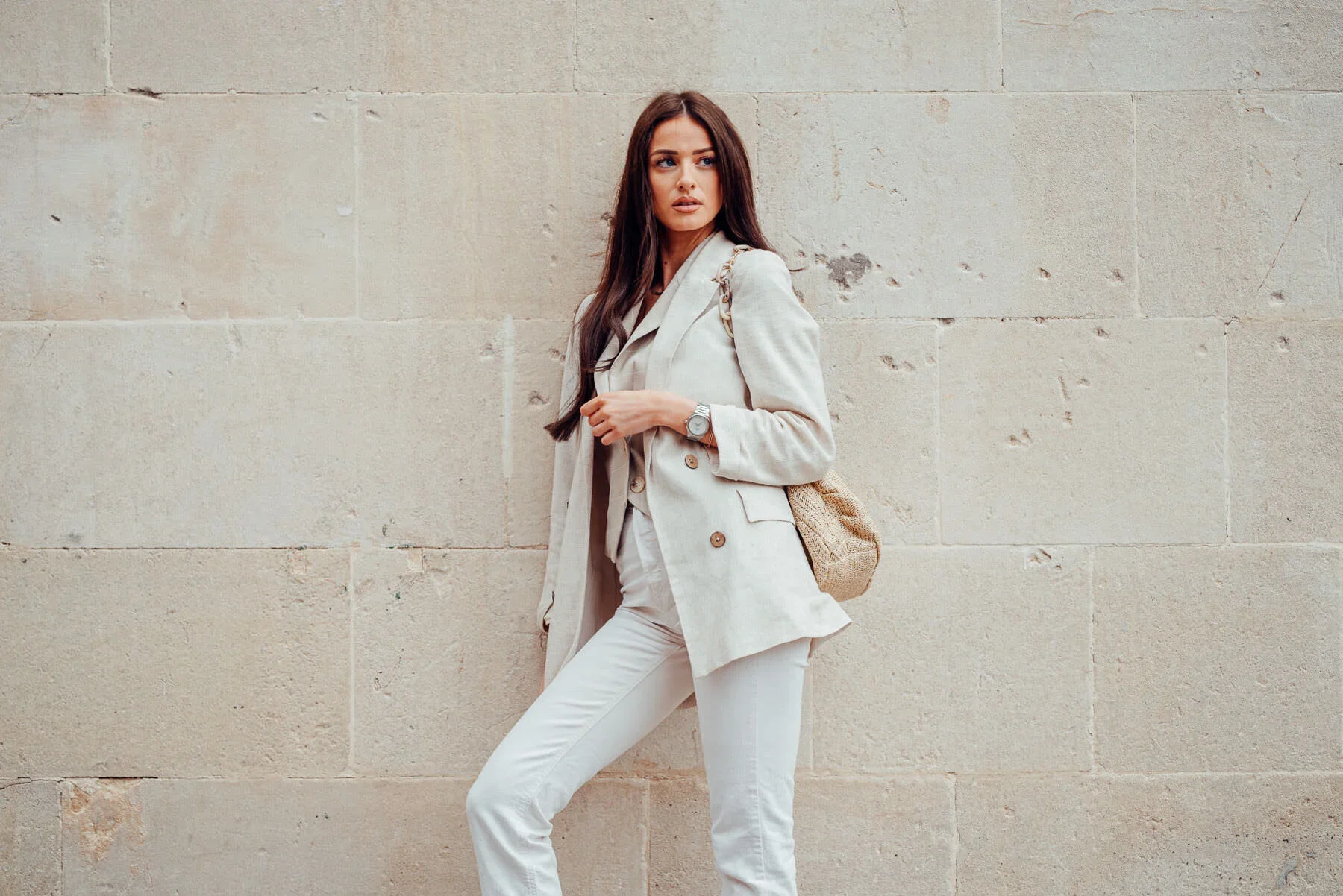 A young woman standing against a beige stone wall, wearing a light-colored blazer, white pants, and a watch, with a beige bag over her shoulder.