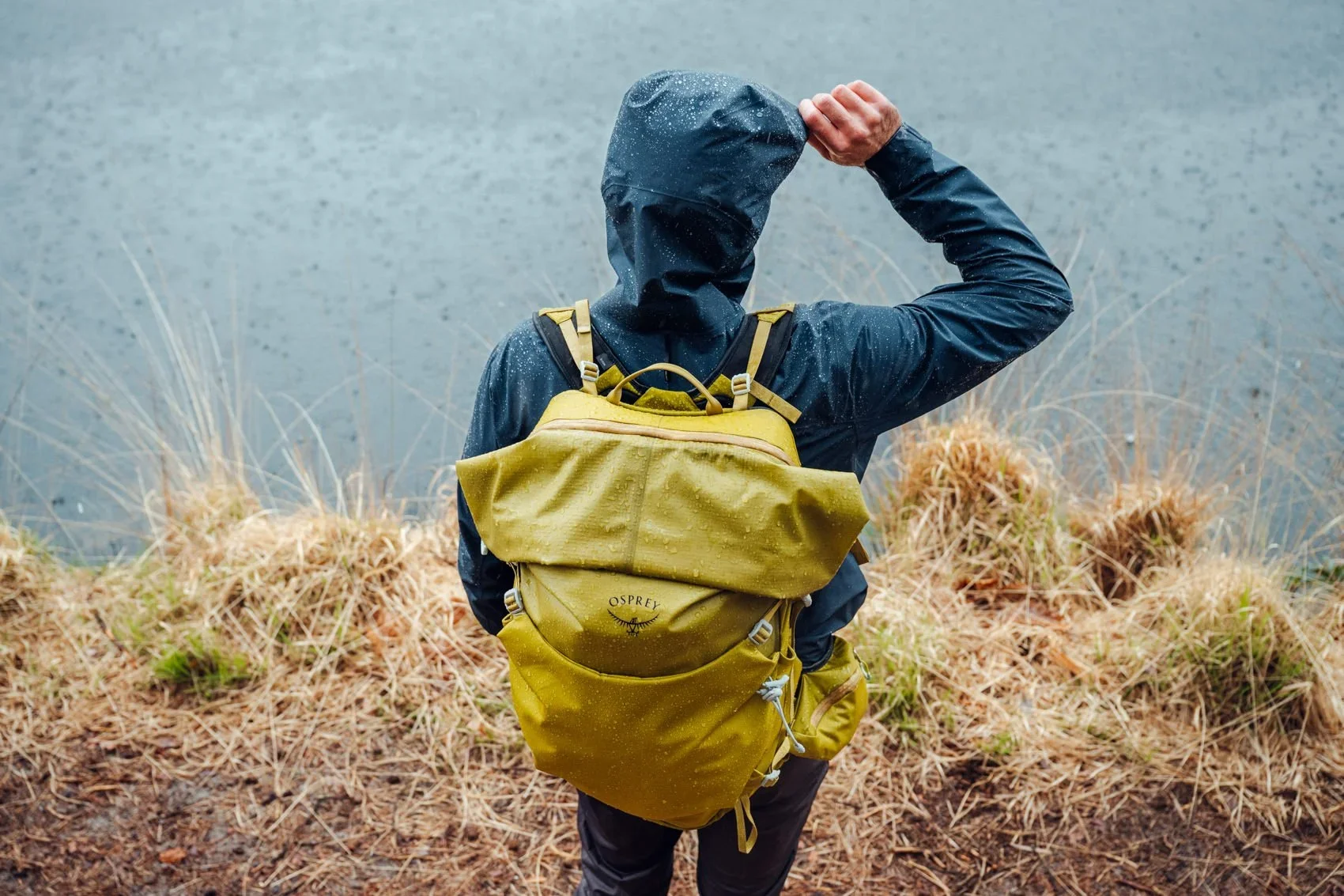 A person wearing a rainy weather jacket and a yellow backpack standing by a body of water, holding their hood with one hand.