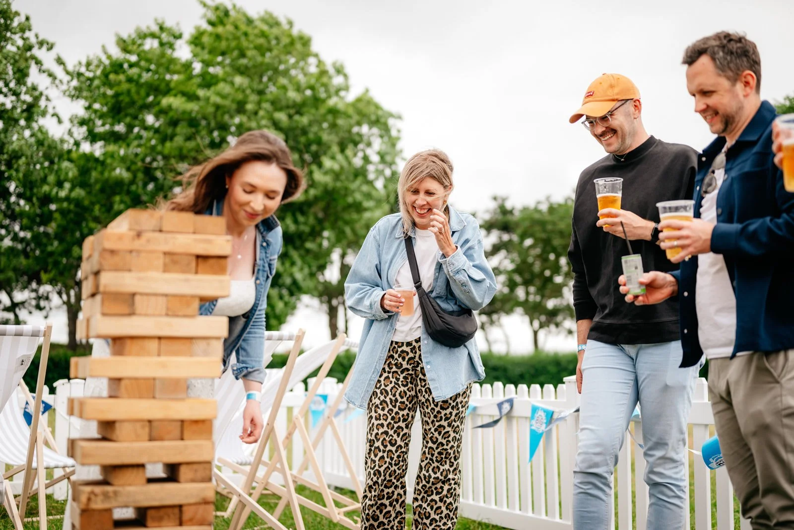 Corporate Summer Event showing a Group of friends playing giant Jenga and drinking beer outdoors at a backyard party with trees and white fence in the background. Photography By James Bridle Event Photographer.