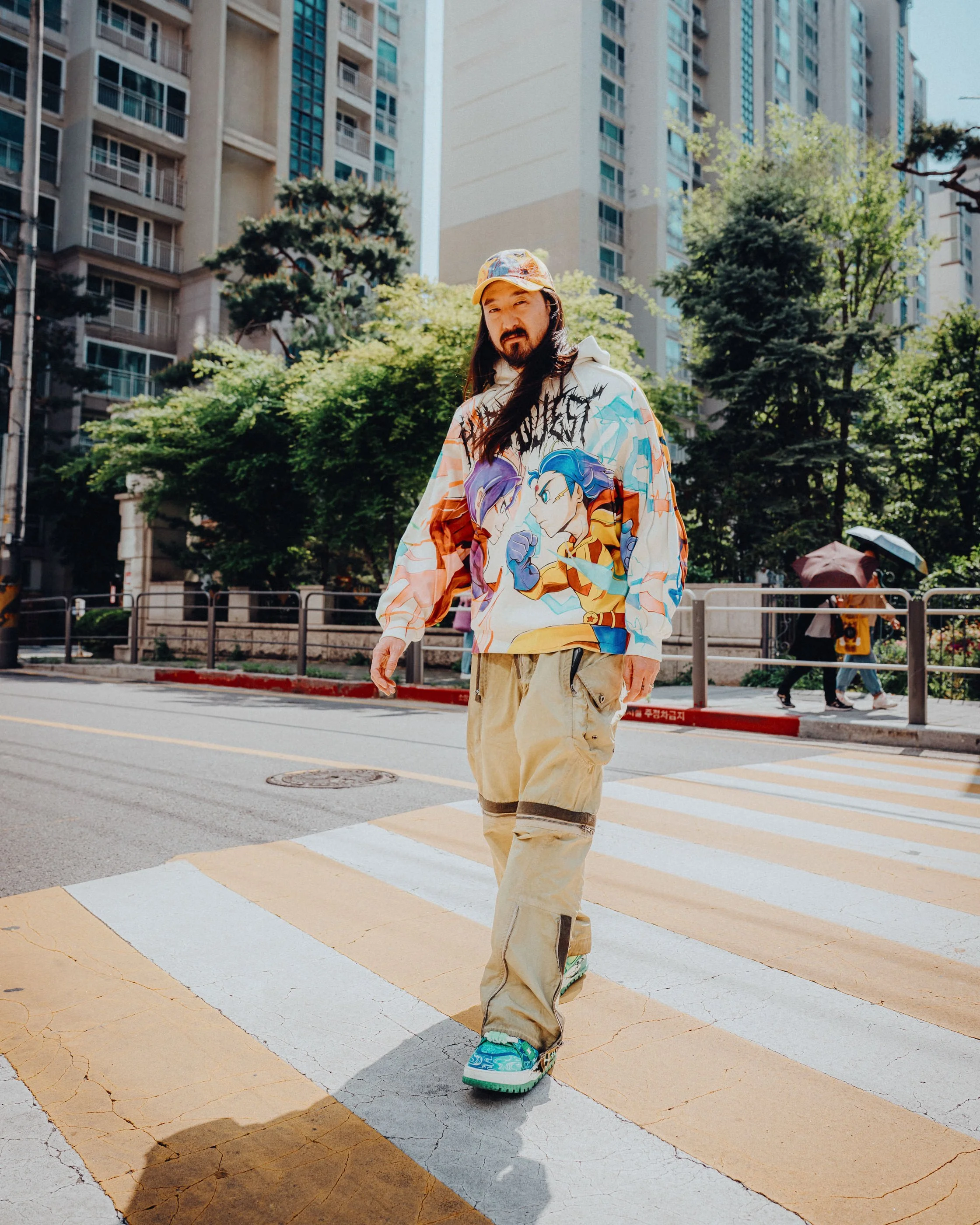 Steve Aoki South Korea by James Bridle Photography. Steve crossing a city street at a crosswalk, wearing a colorful hoodie with anime characters, beige pants, and bright sneakers, with tall buildings and trees in the background.