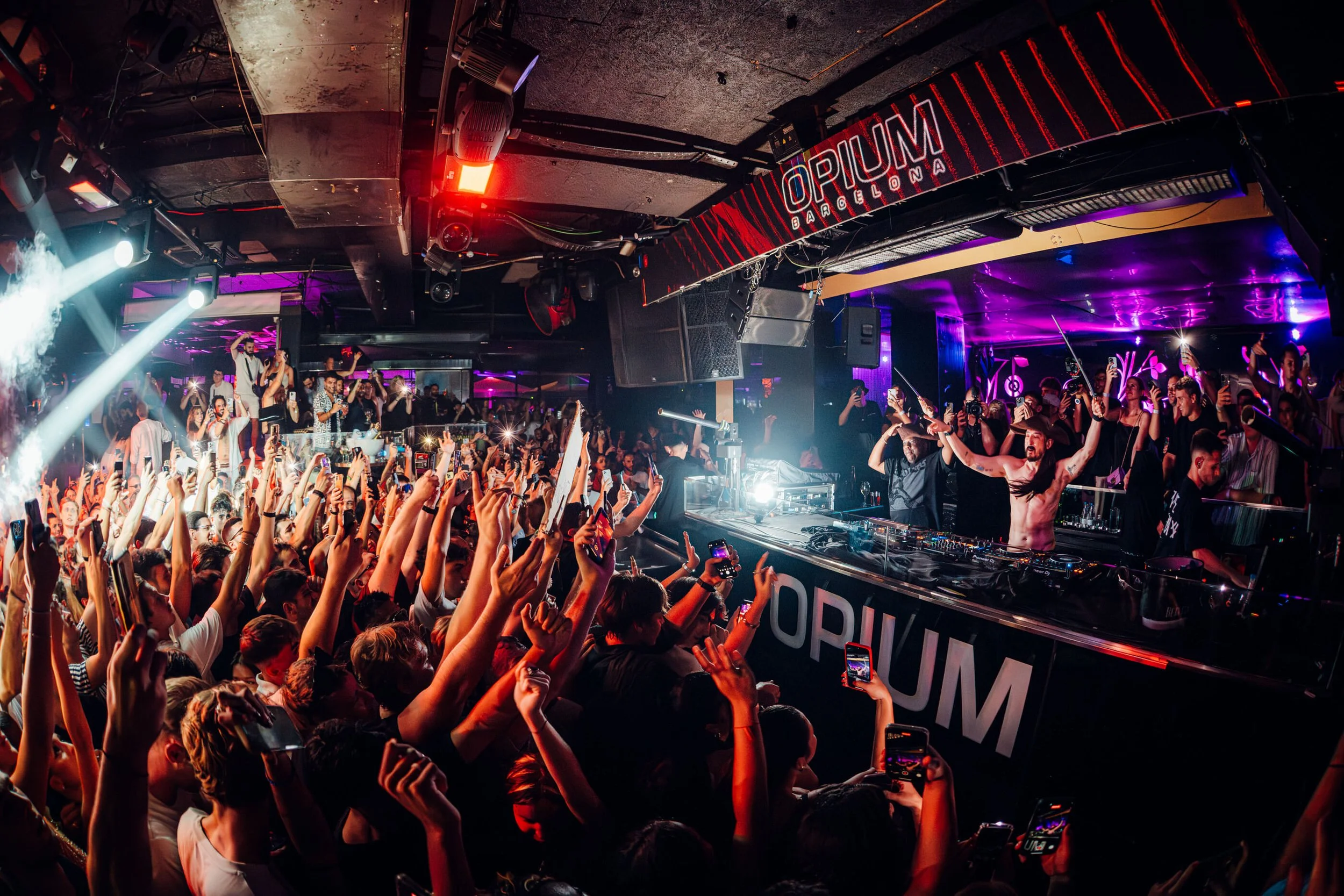 Steve Aoki plays in Opium in Barcelona surrounded by enthusiastic fans raising their hands and taking photos. The club has vibrant purple and blue lighting, with a sign that reads 'Opium Barcelona'.