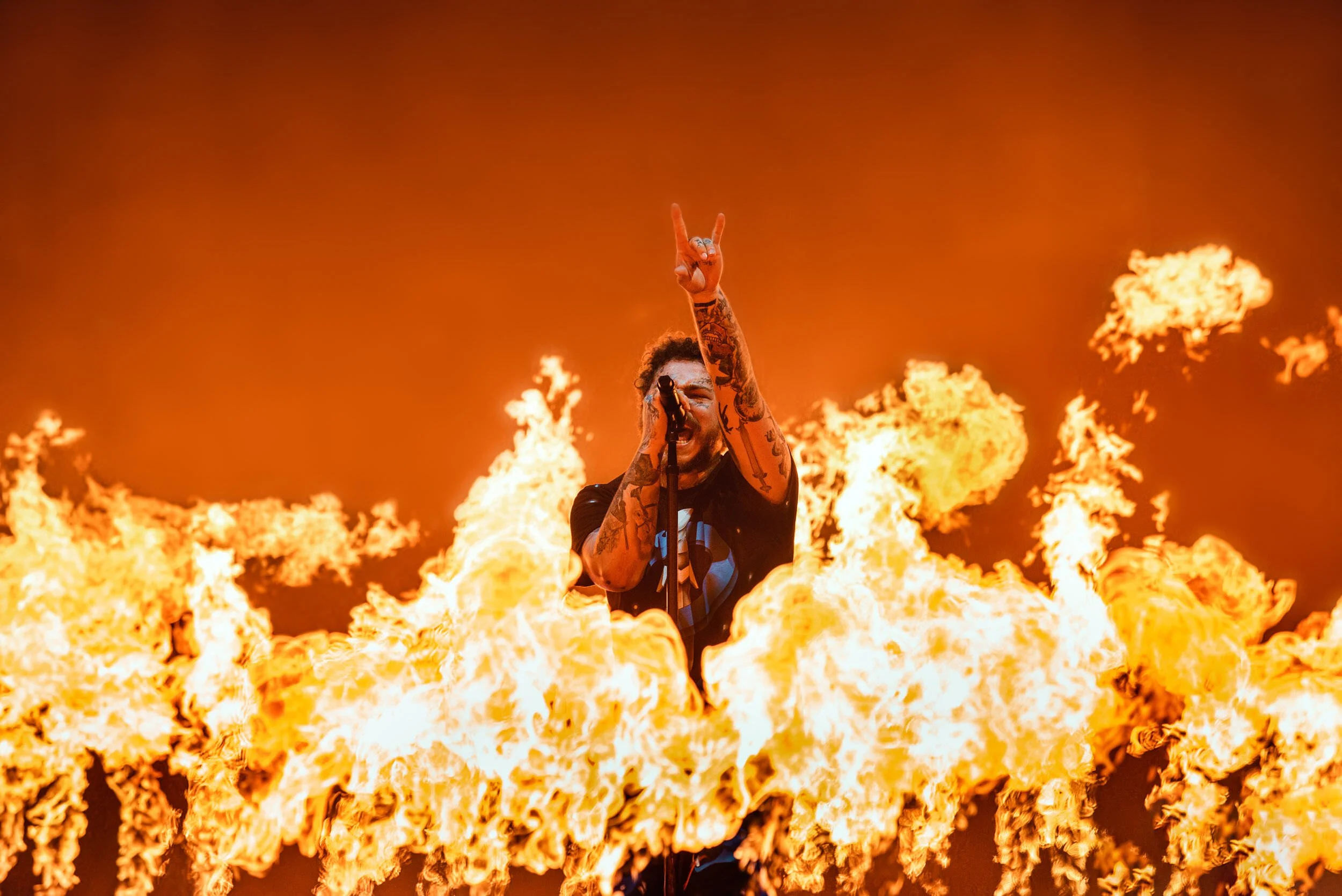 Post Malone at Reading Festival 2019 singing or shouting into a microphone, surrounded by intense flames on stage during a performance.