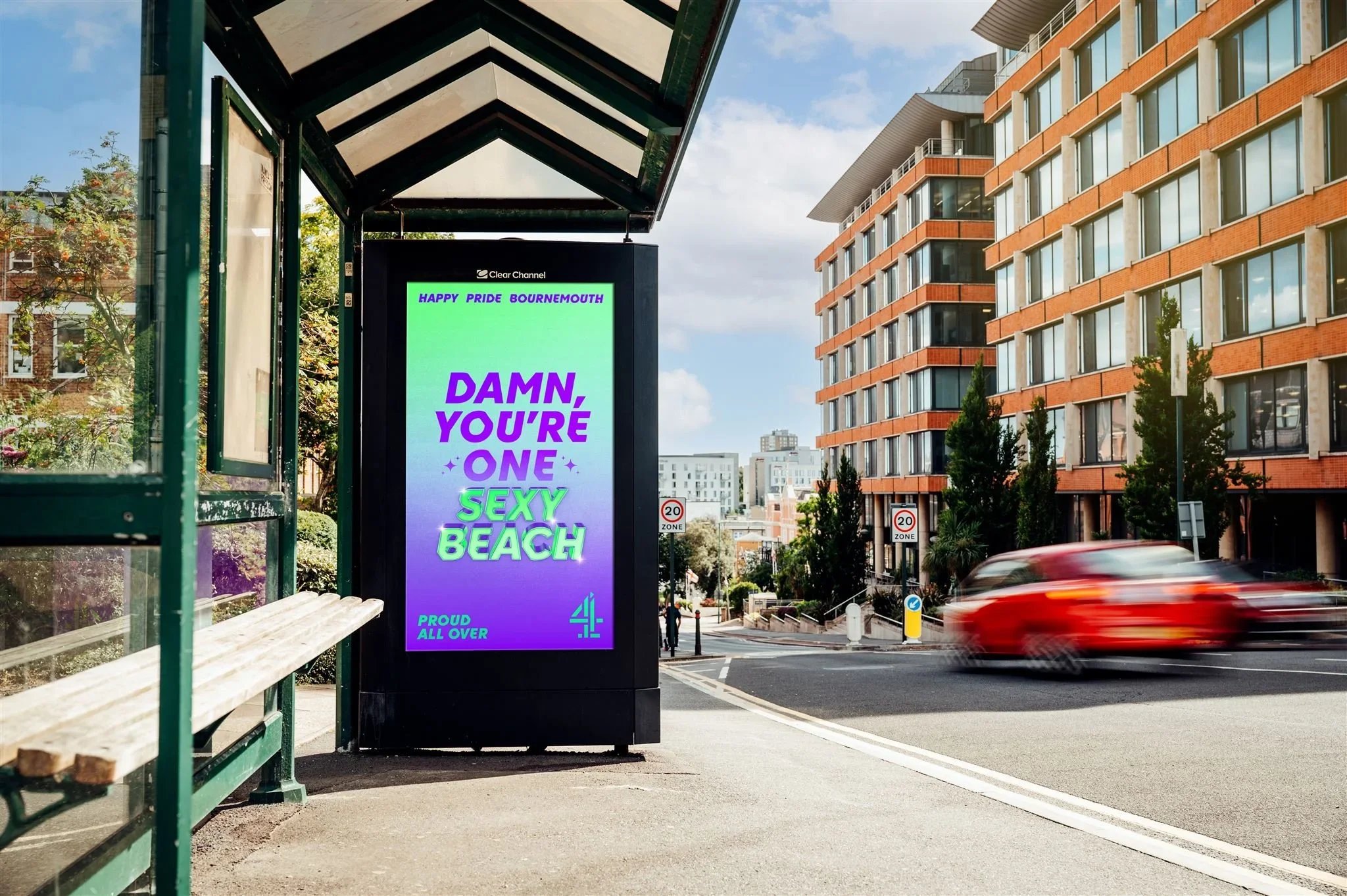 Digital billboard at a bus stop displaying a colorful message for Bournemouth pride, reading, 'Damn, you're one sexy beach,' with smaller text above saying 'Happy Pride Bournemouth' and below 'Proud all over,' next to a sidewalk and a blurred red car