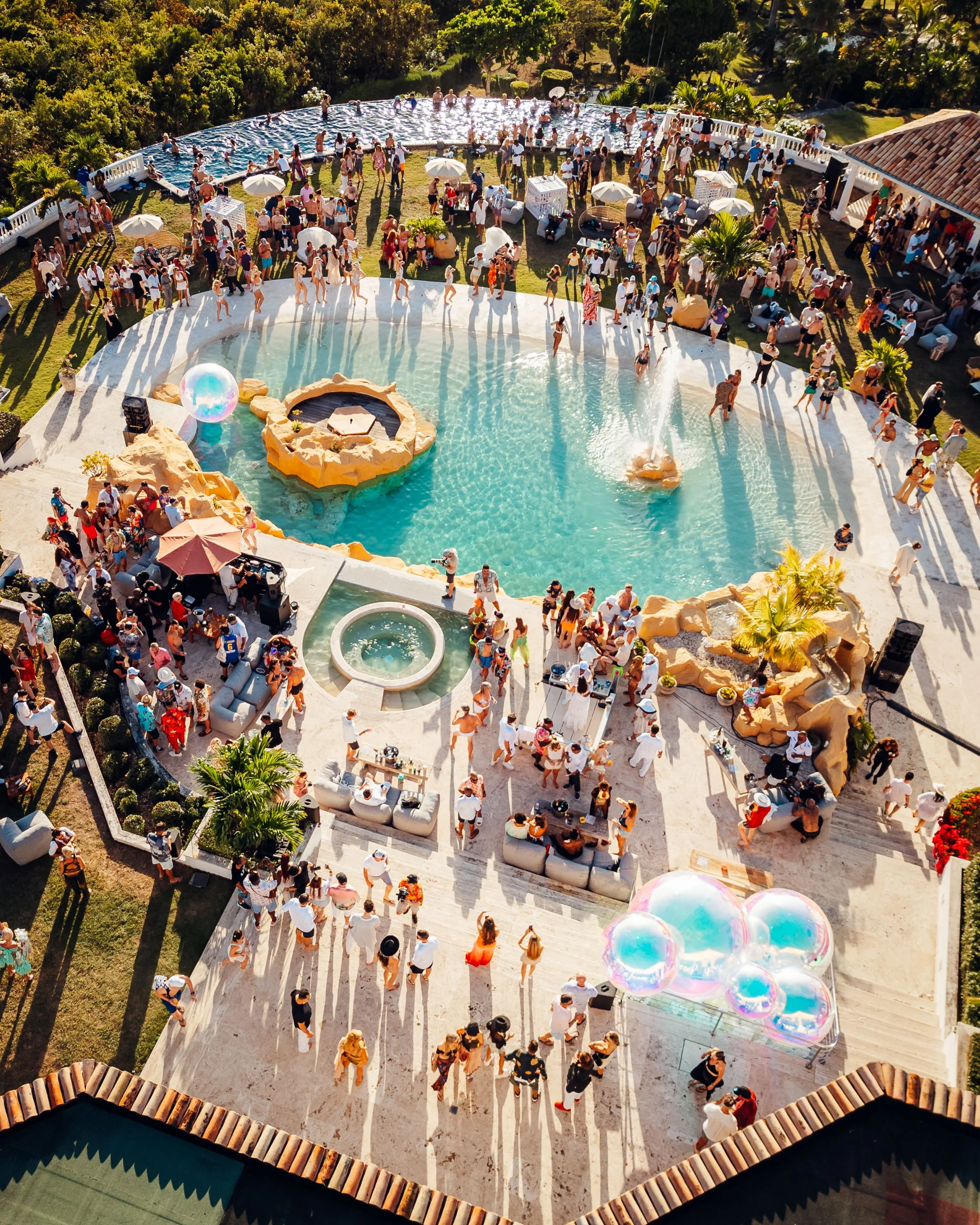 SXM Festival VIP Villa Party socialising on lounge chairs and around the pool, with umbrellas and decorative elements like balloons and palm trees. Captured by James Bridle