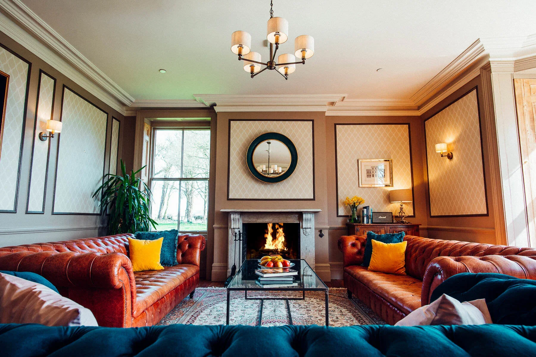 Abbots Court Rural Retreat showing the living room with two brown leather sofas, a glass coffee table, and a fireplace with a fire burning. The room has beige panelled walls, a chandelier, and wall sconces, with a large window showing trees outside.