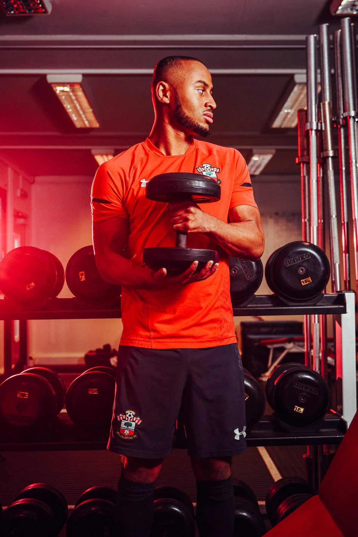 A man in a red sports shirt and black shorts holding a dumbbell in a gym with weight plates on racks behind him.
