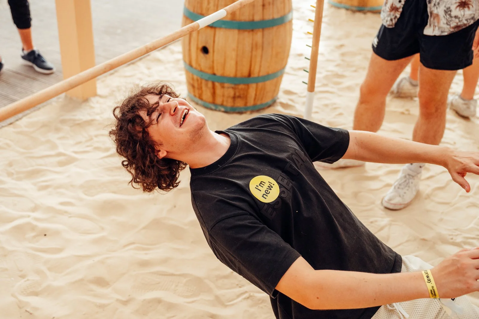 Corporate Summer Event in London showing A young man with curly hair lying on the sand, smiling and pointing, wearing a black t-shirt with a yellow sticker that reads 'I'm new!'. There are people in the background, and a barrel with a pole nearby. 