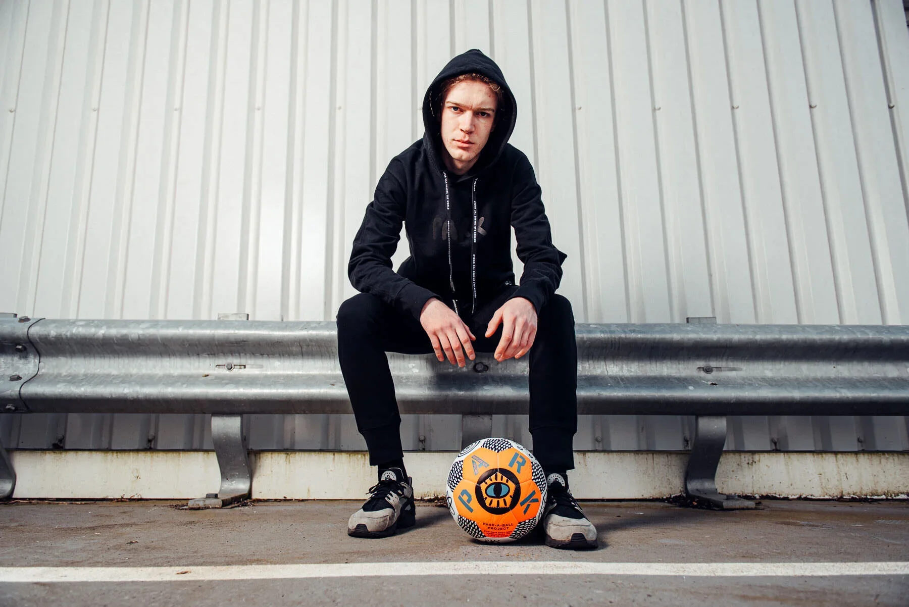 Sports Fashion Shoot by James Bridle. A young man dressed in black sitting on a metal bench outdoors with a soccer ball placed on the ground in front of him. He is wearing a hoodie with the hood up, dark pants, and sports shoes. 