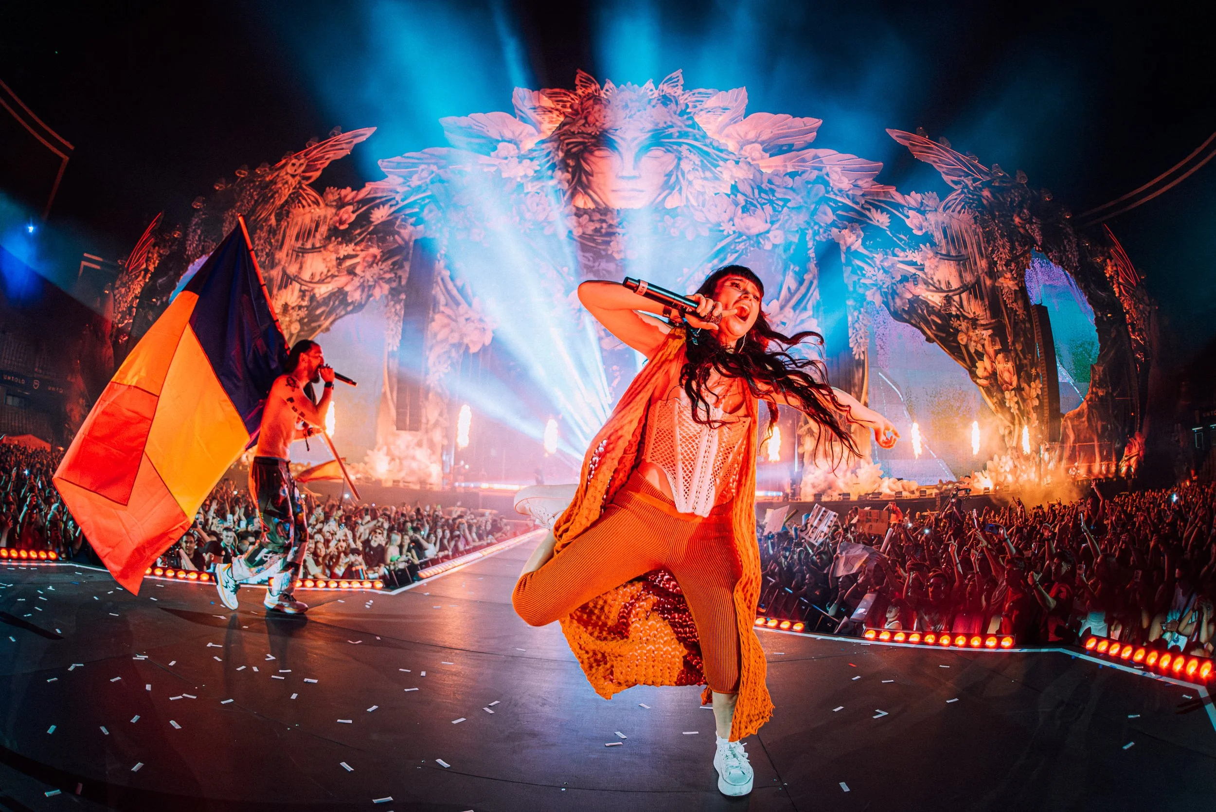 Steve Aoki on stage at UNTOLD Romania holding a microphone and a flag of the country with a large animated face and flames in the background, and a crowded audience taken in 2024 by James Bridle Music Photographer. 