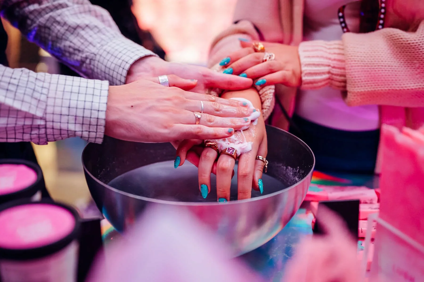 LUSH Oxford Street. Multiple people with colorful manicured nails are washing their hands or nails in a bowl of water with soap, surrounded by makeup products.