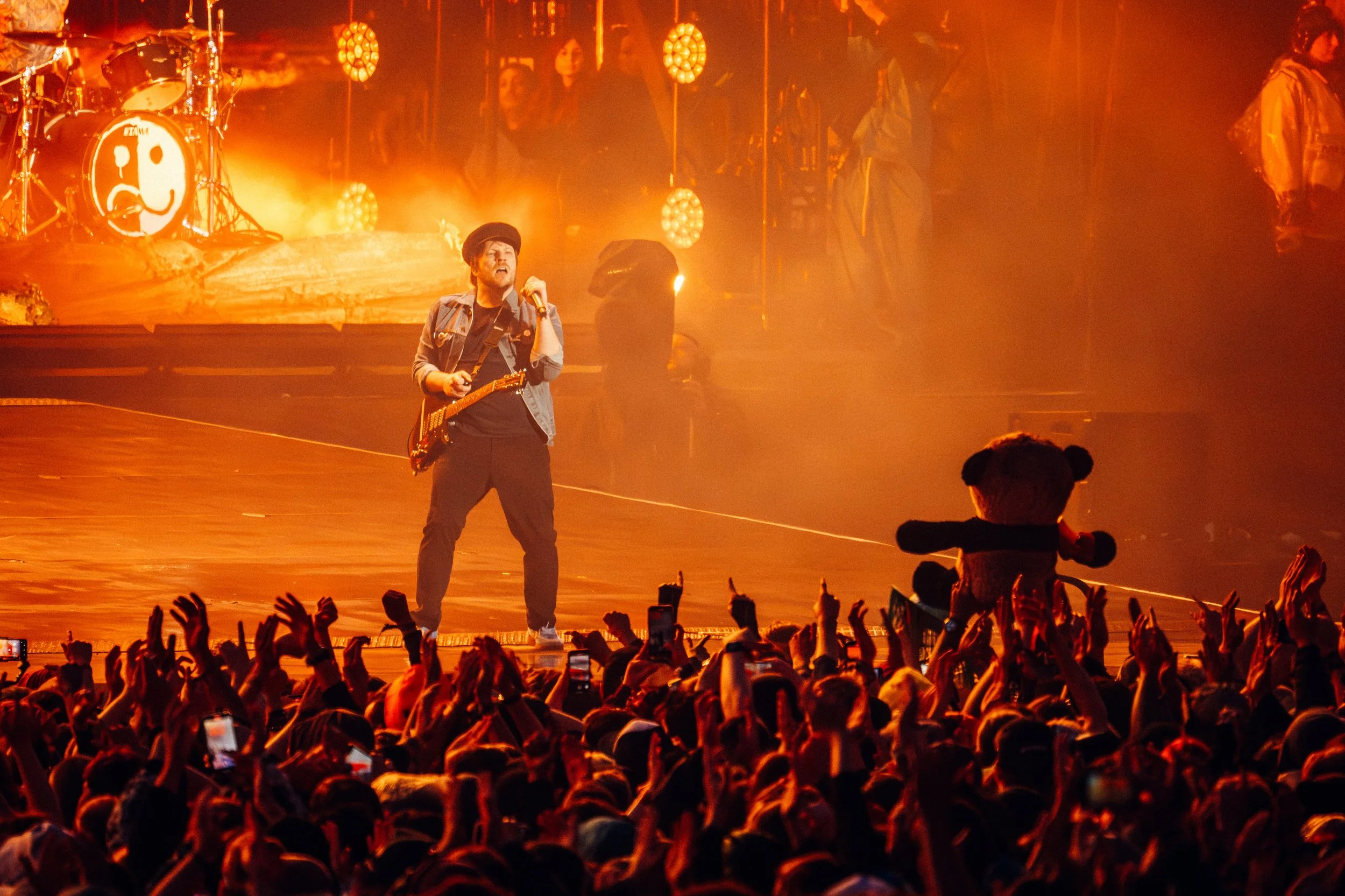 Fall Out Boy's - Patrick Stump standing on stage in front of a large crowd. The stage is illuminated with warm, orange lighting, and there is a drummer's set in the background with a smiley face on the bass drum. 