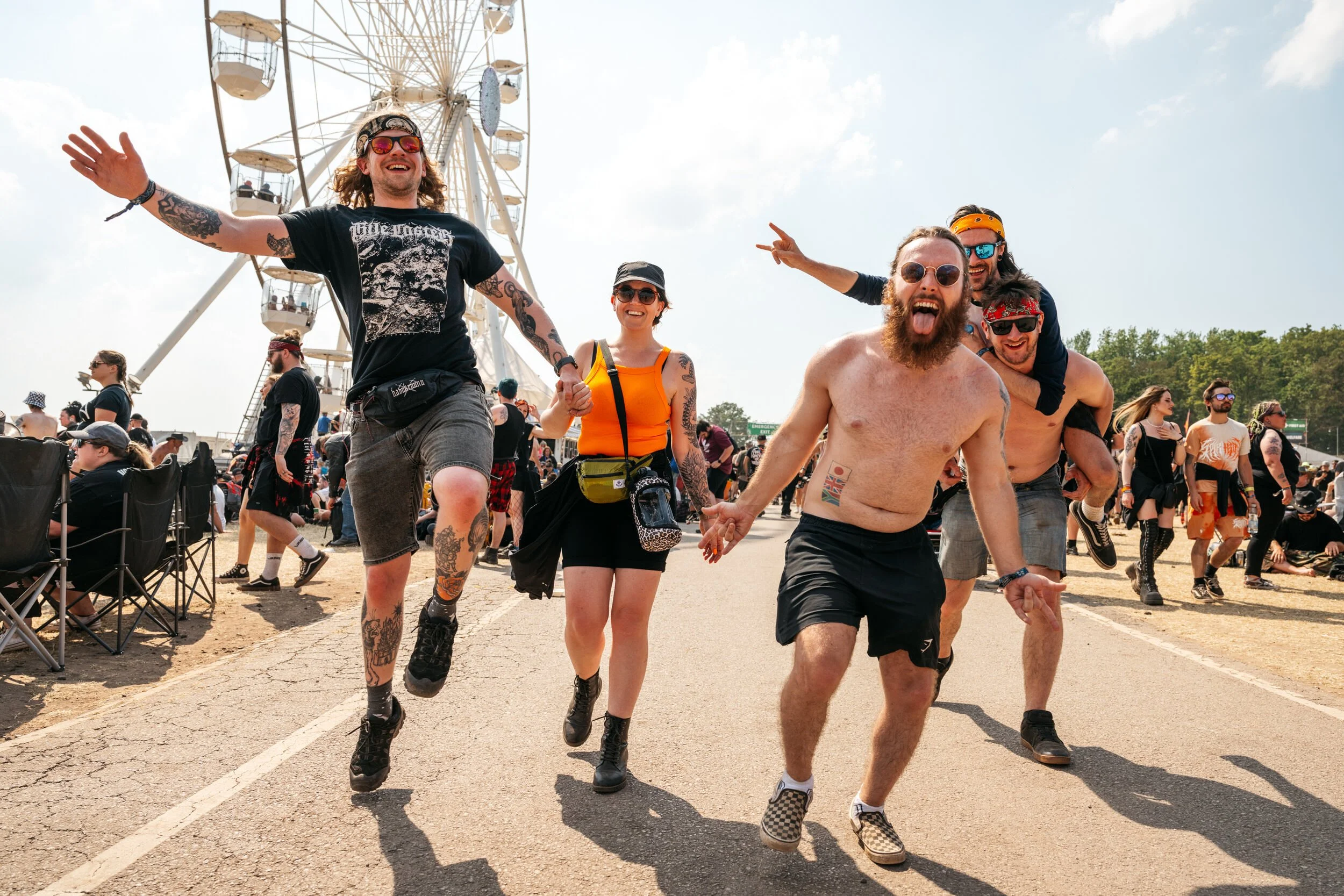 Download Festival 2024 - Group of friends at an outdoor festival or fair, walking and dancing on a dirt path near a Ferris wheel, with some people sitting and others standing in the background, enjoying sunny weather.
