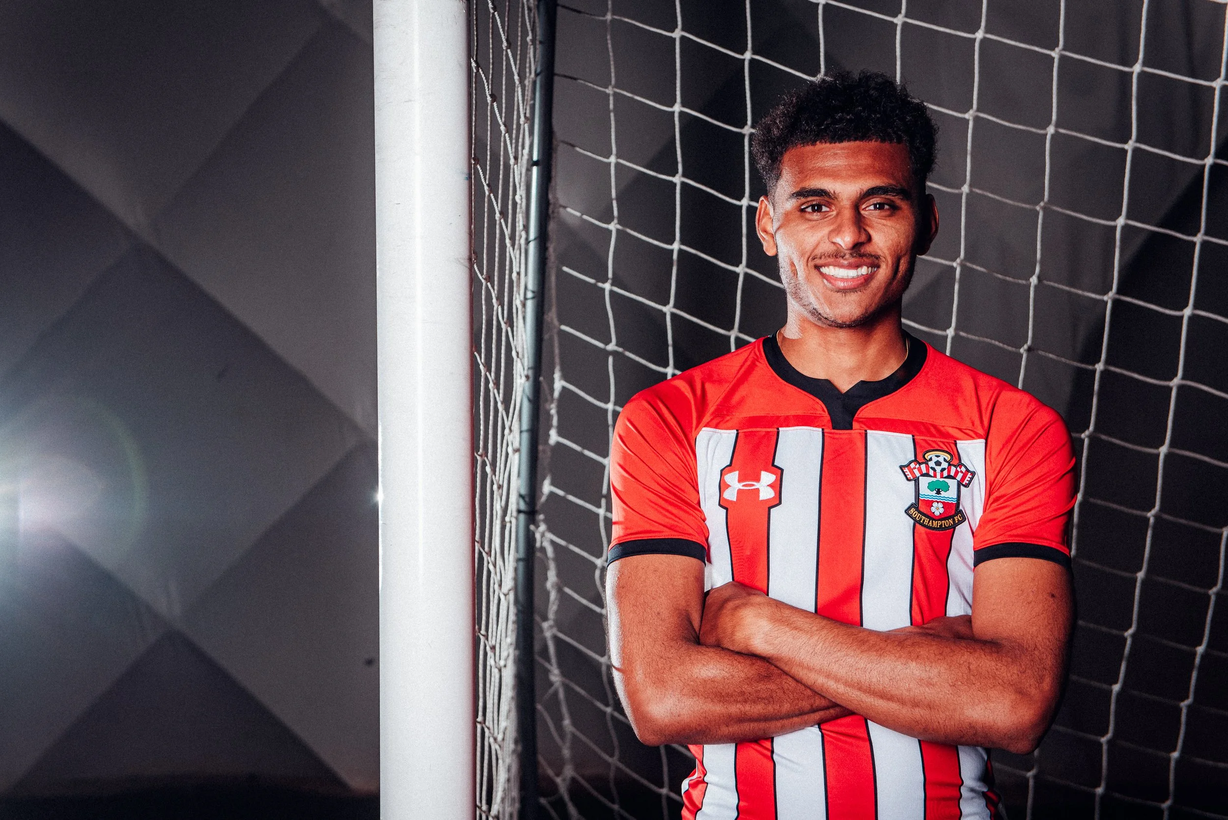 Marcus Barnes during a player shoot by James Bridle,  seen in a red and white striped Southampton FC football jersey standing with arms crossed, smiling in front of a goal net on a dark background.