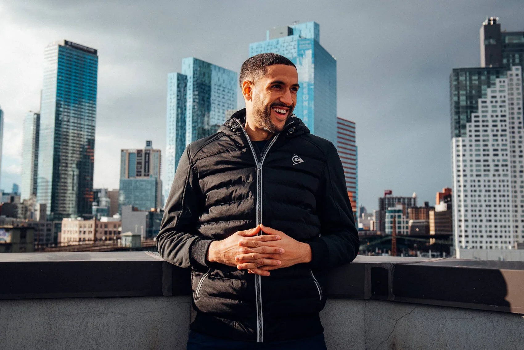 A man smiling and standing on a rooftop with a city skyline in the background, wearing a black jacket.