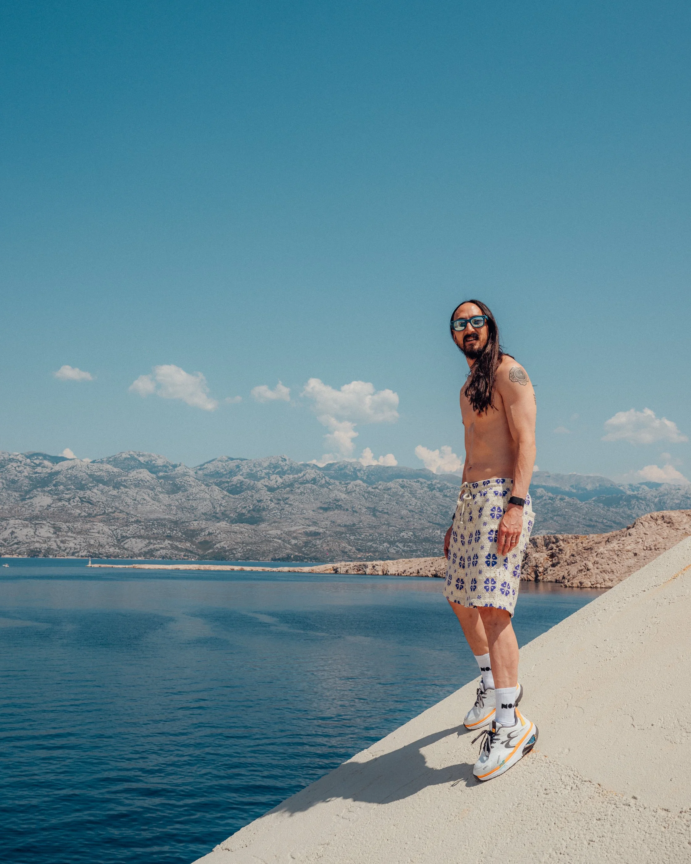 Steve Aoki in Croatia standing on a sloped concrete surface near water with mountains in the background under a clear blue sky.
