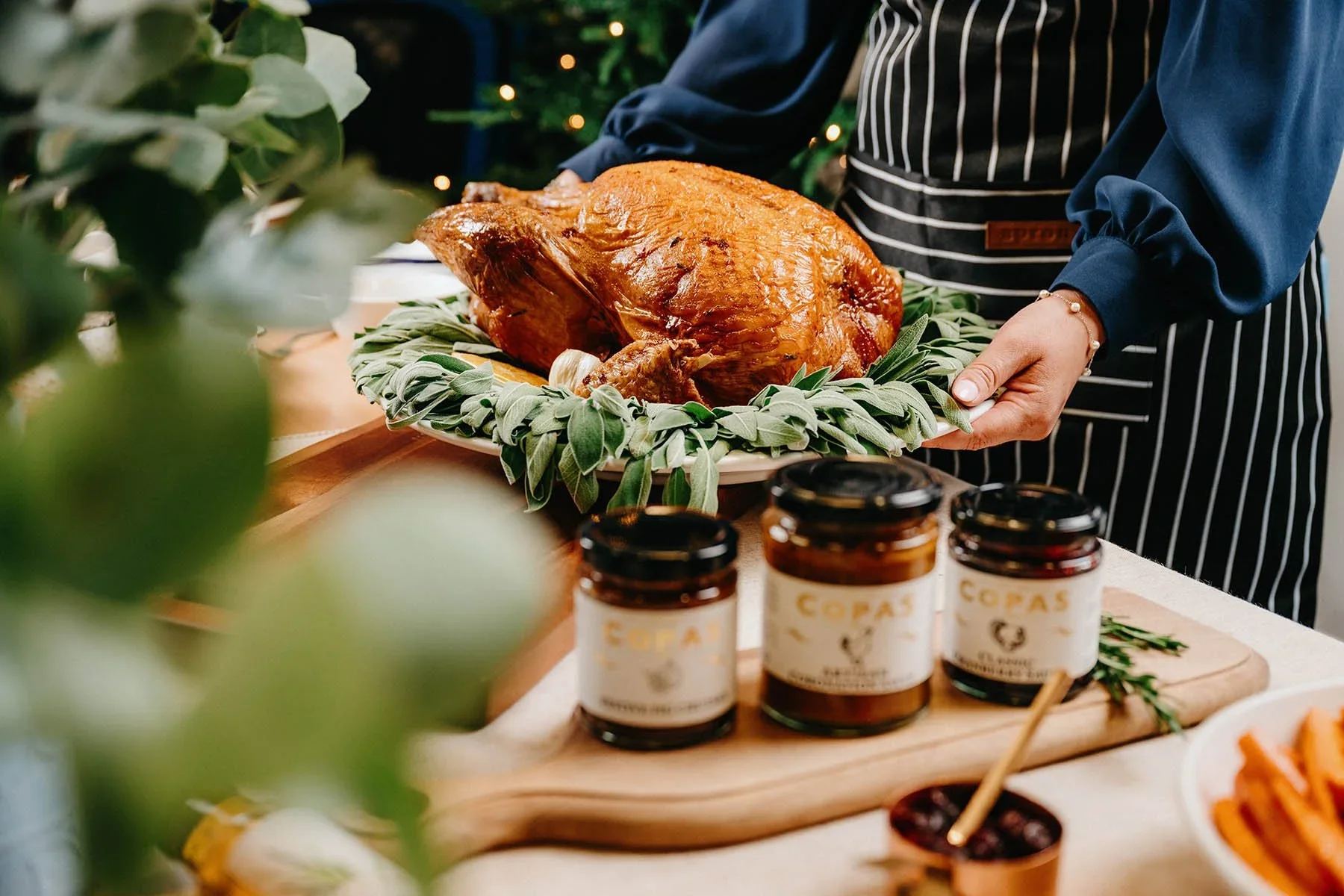 Person holding a tray with a roasted turkey garnished with herbs at a festive gathering.
