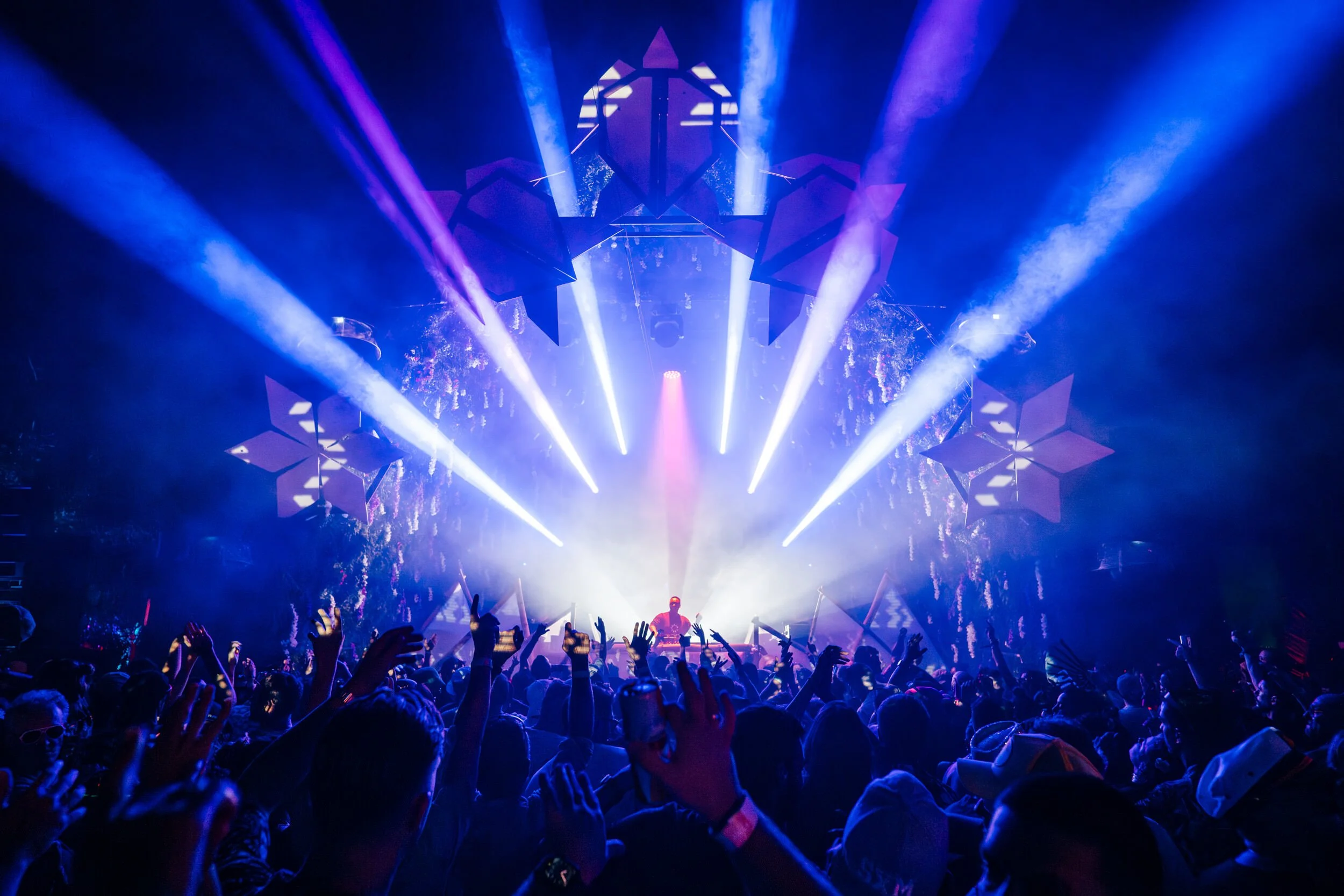 SXM Festival showing Happy Bay Arena - with a DJ performing on stage surrounded by vibrant blue and purple lights, with an enthusiastic crowd raising their hands. Captured by James Bridle Music Photographer in 2024