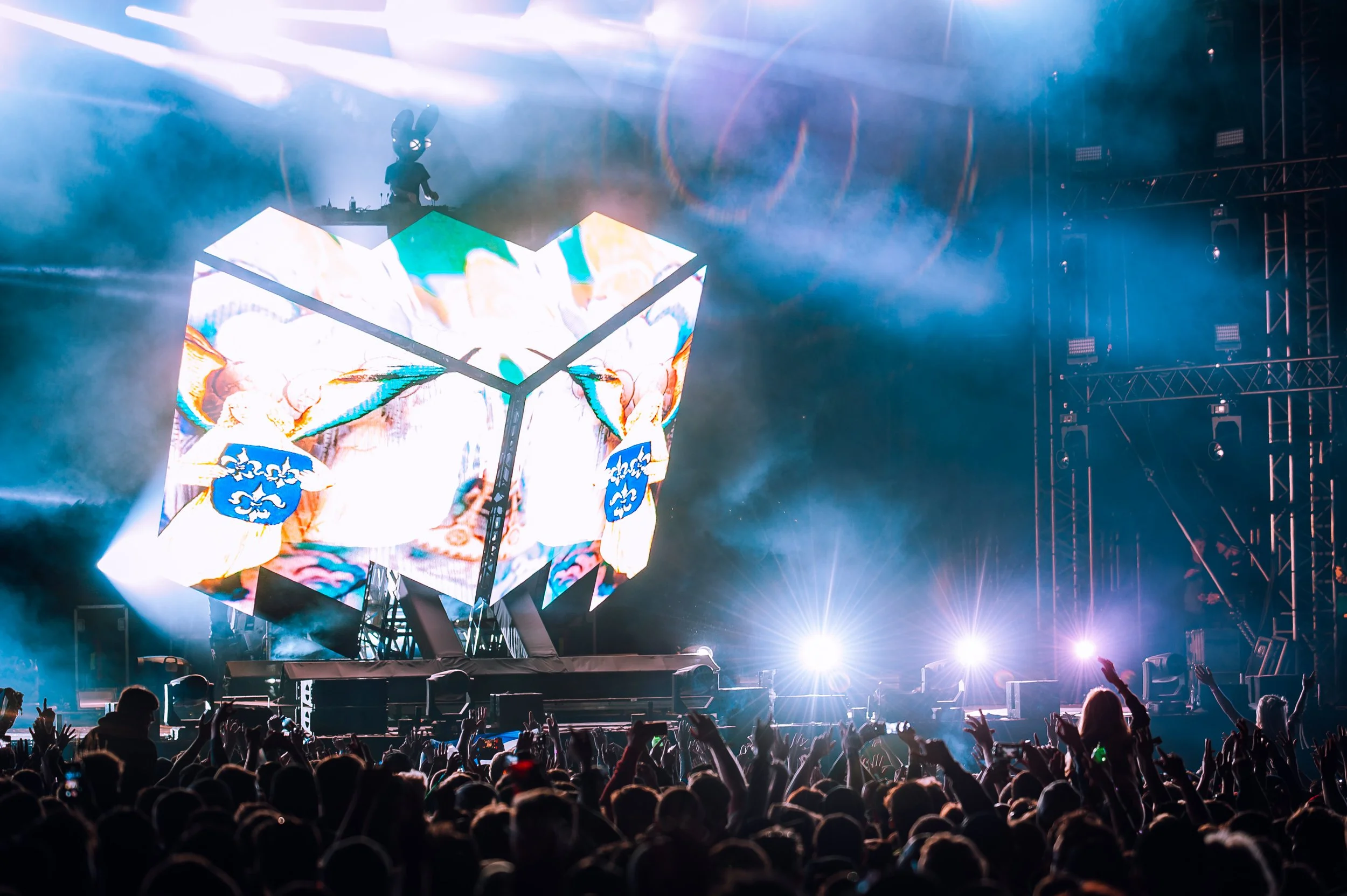 DeadMau5 at Creamfields 2017 performs on a stage with a large LED screen showing colorful visuals, while a crowd of people raises their hands in front of the stage at a concert or music festival.