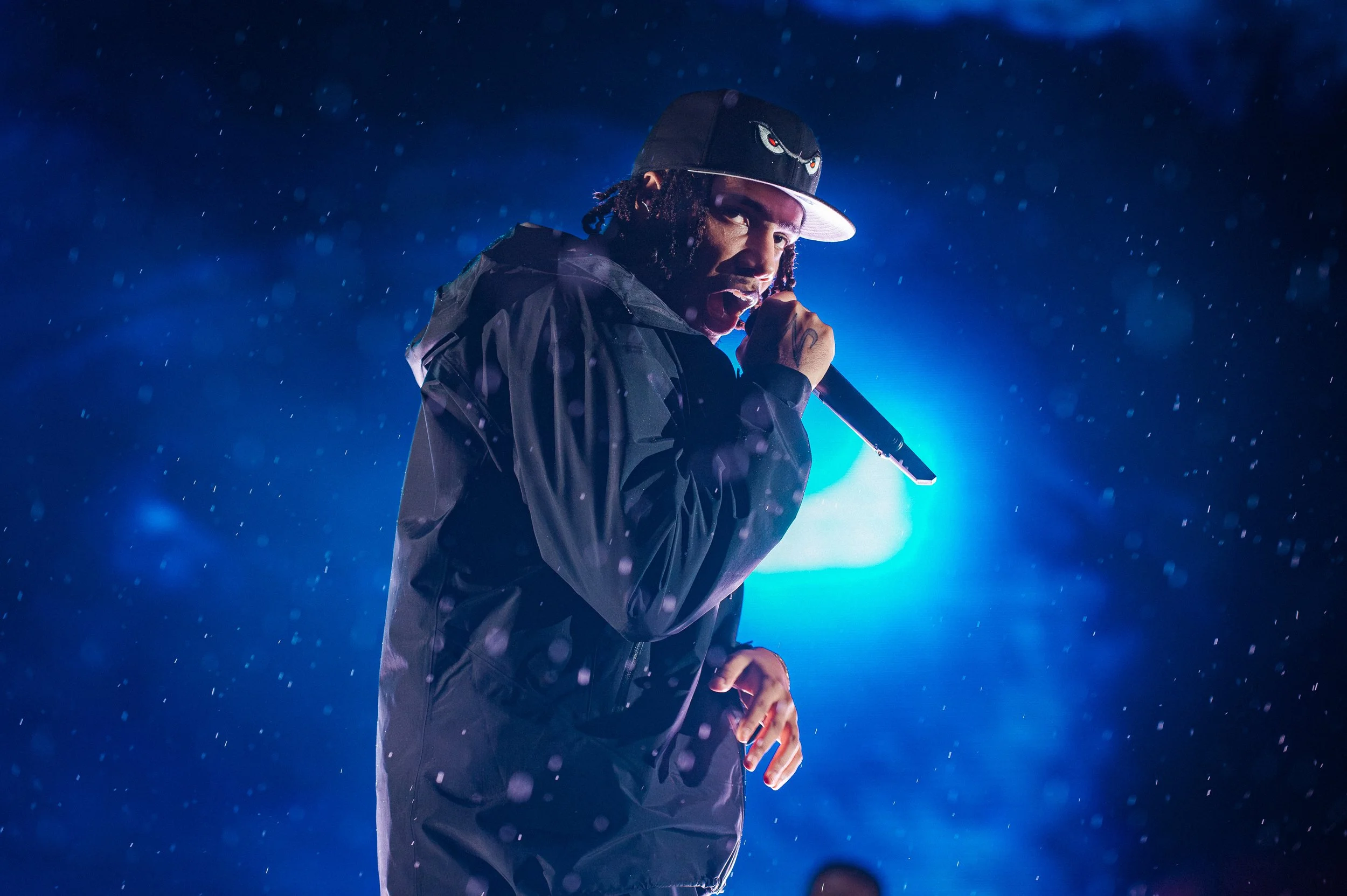 AJ Tracy at Parklife Festival 2021 captured by James Bridle holding a microphone near his mouth, wearing a black jacket and a black cap with a logo, backlit by blue stage lights.