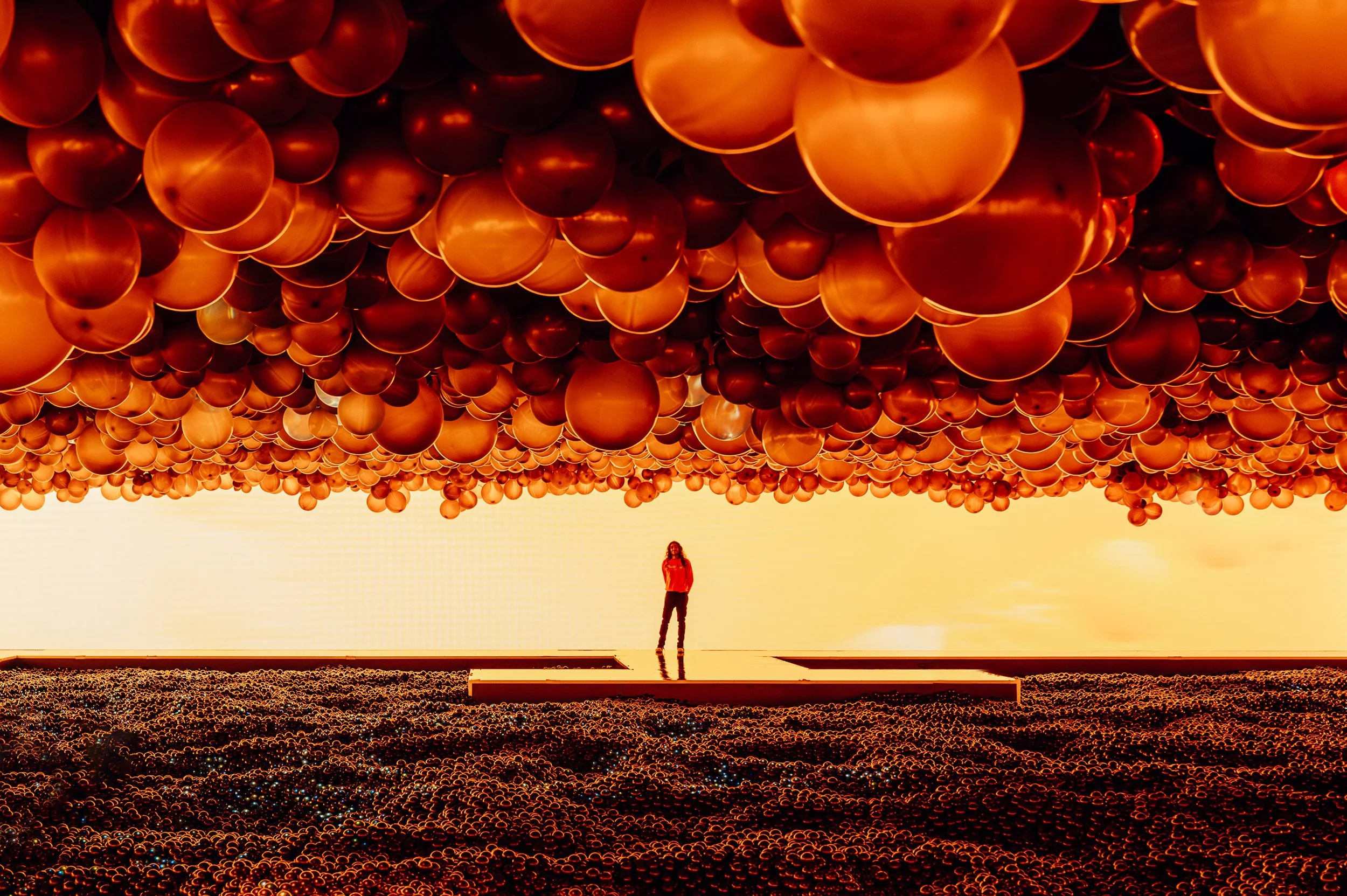 NYC Balloon Event - A person standing inside a large installation of hanging copper-colored balloons creating a ceiling, with a ground surface also covered in similar balloons, illuminated with warm, golden lighting.