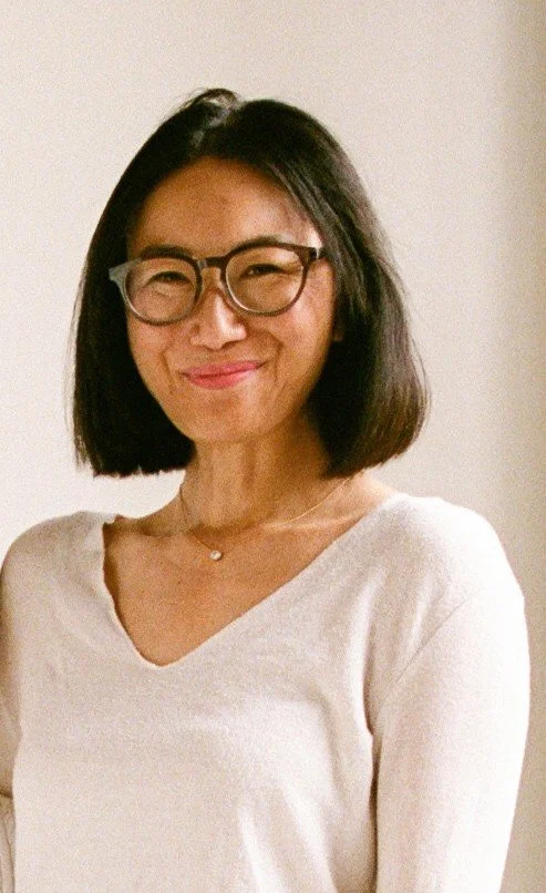 A woman with glasses and shoulder-length black hair smiling, wearing a white V-neck top and a necklace.
