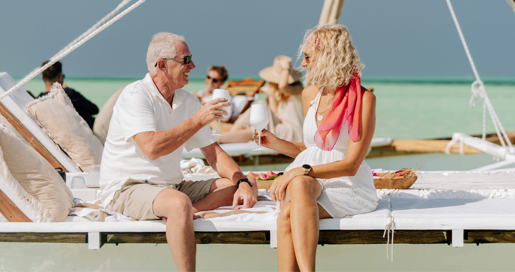 A man and a woman sitting on a boat, enjoying drinks and smiling at each other, with a beach and other people in the background.