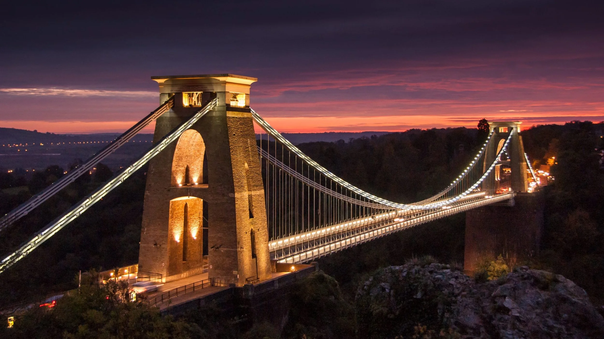 A suspension bridge illuminated at night with a colorful sunset sky in the background.