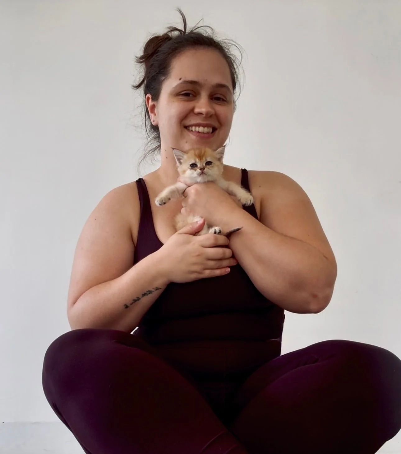 A woman sitting cross-legged and smiling at the camera while holding a small ginger kitten against her chest.