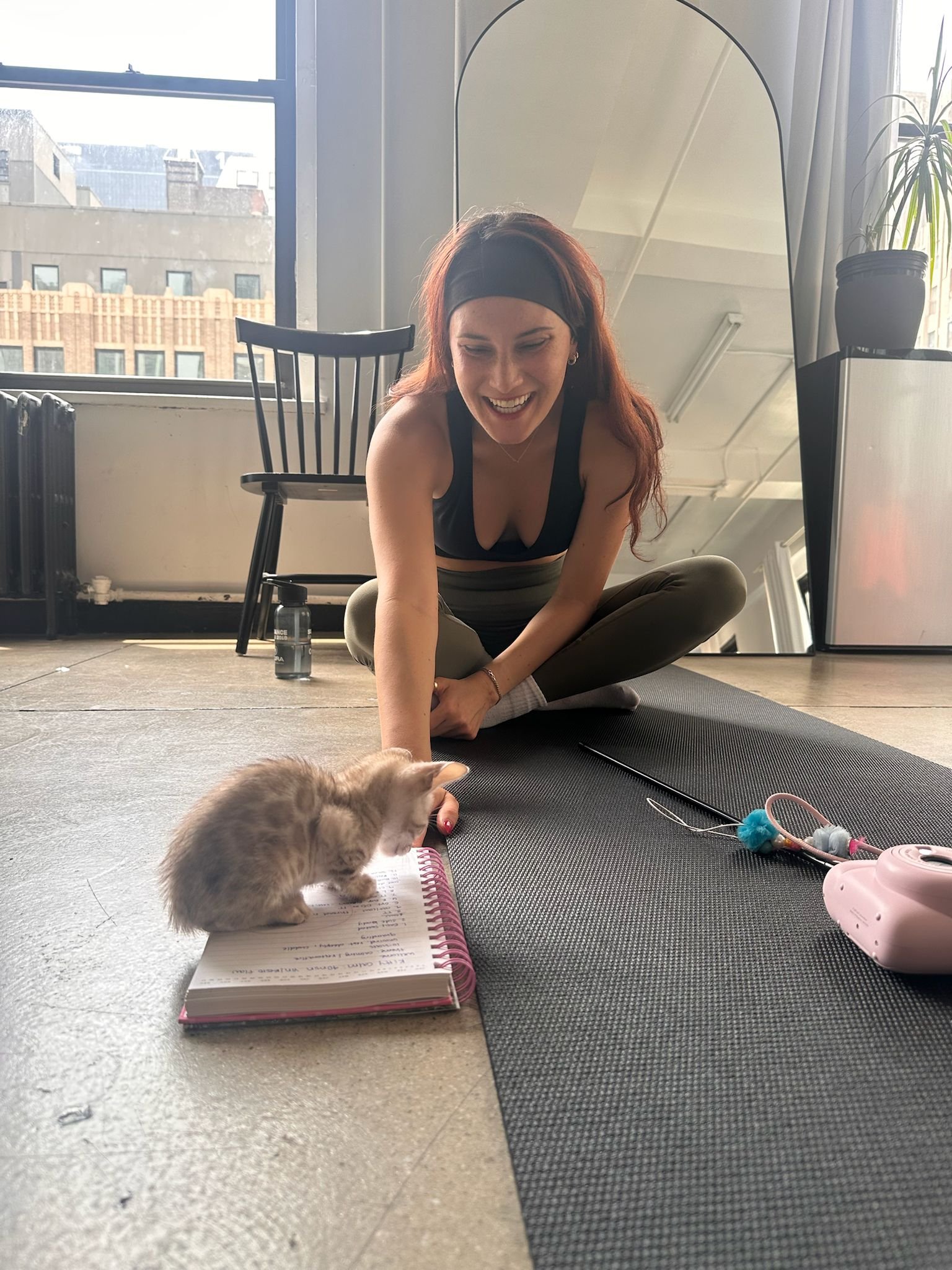 A woman with red hair, wearing a black headband and sports bra, sitting cross-legged on a yoga mat, smiling and playing with a small kitten on a notebook, next to cat toys and a pink toy camera, in a bright room with large window and city view.